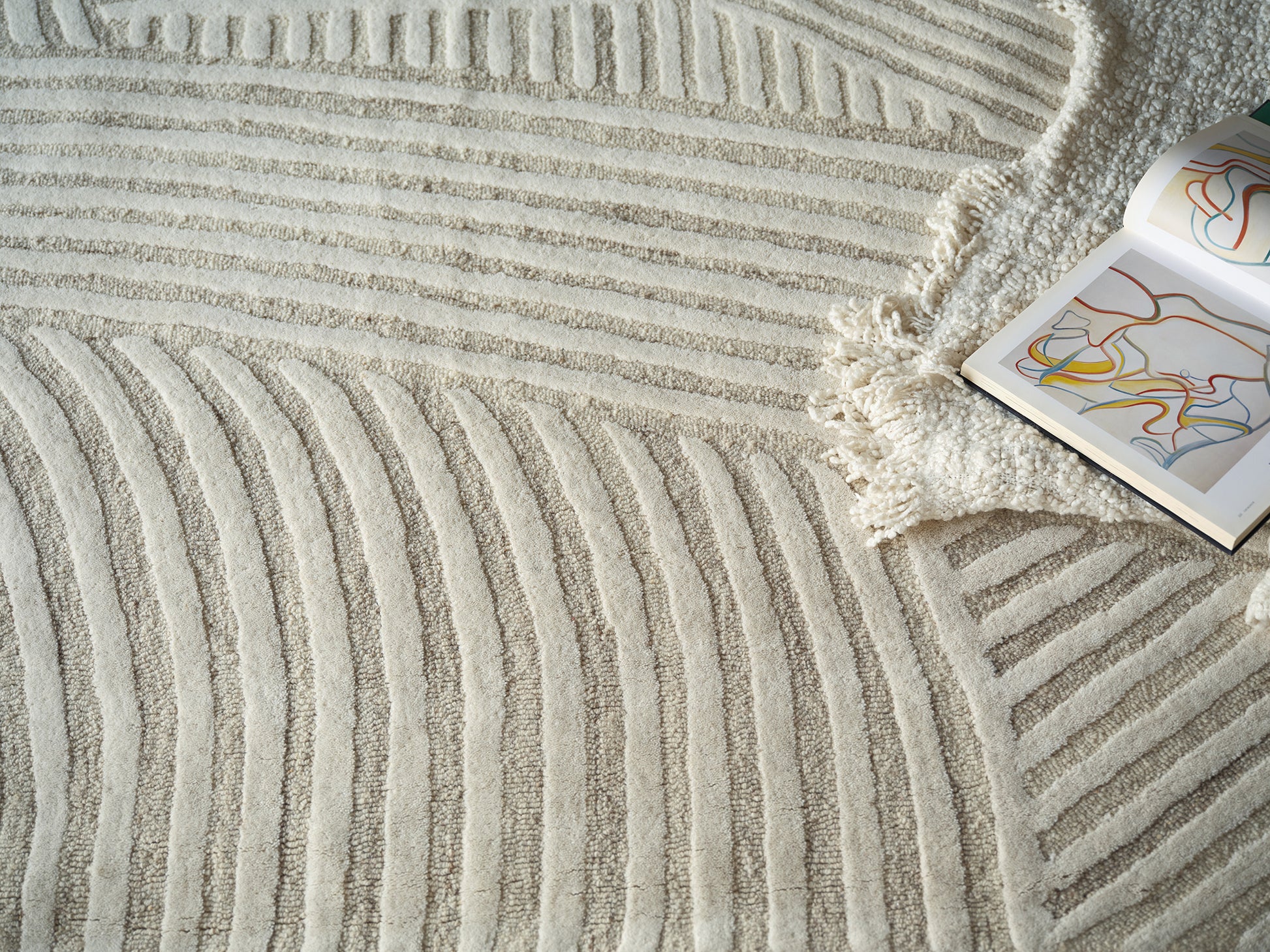Cream shag rug with raised curved stripe pattern and fringe, beside an open art book with colorful abstract lines.