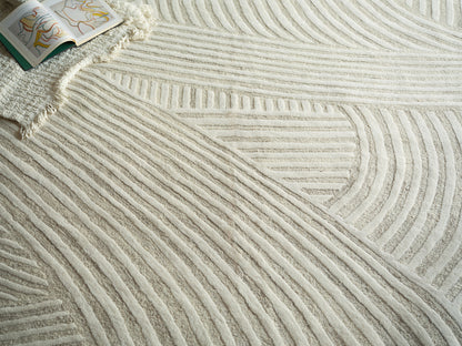 Ivory textured area rug with raised linear stripes and curved pattern; open colorful art book on fringed edge.