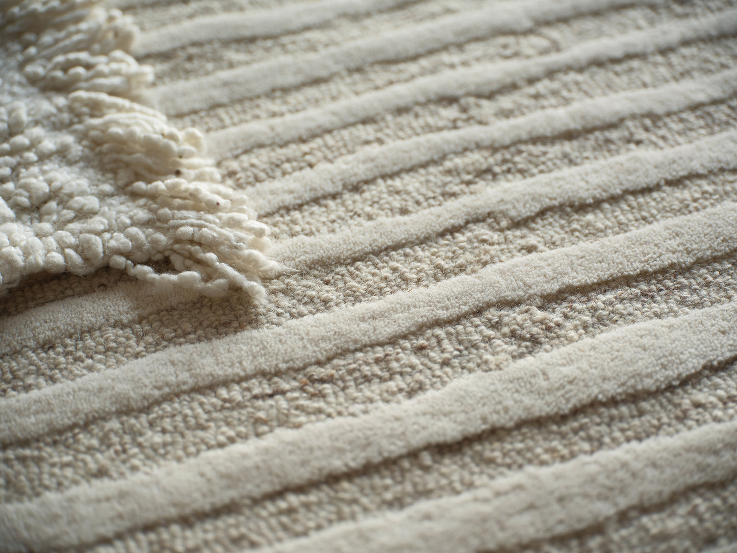 Beige textured rug with ribbed stripes and a cream fluffy fringe along the edge.