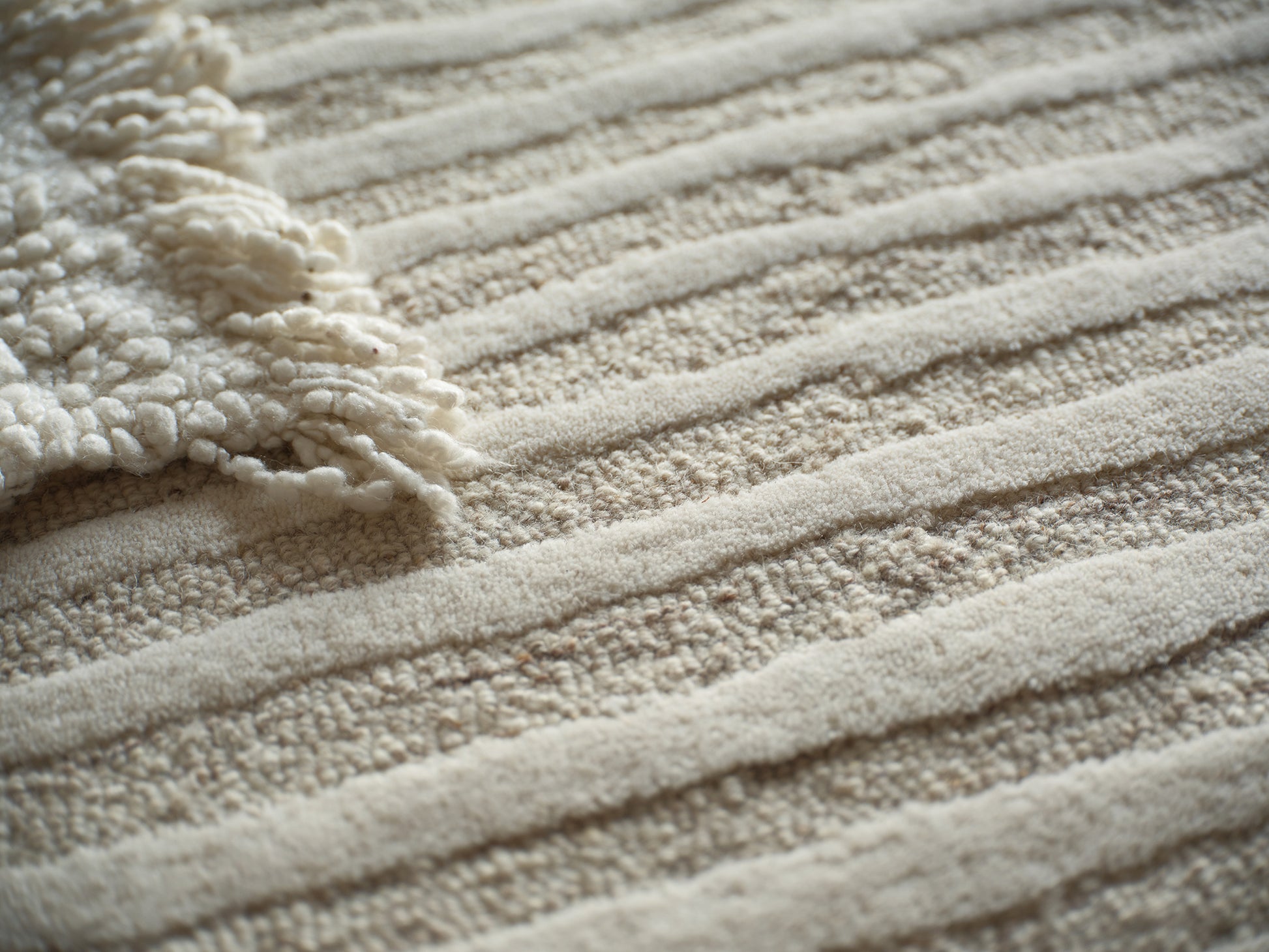 Beige textured rug with ribbed stripes and a cream fluffy fringe along the edge.