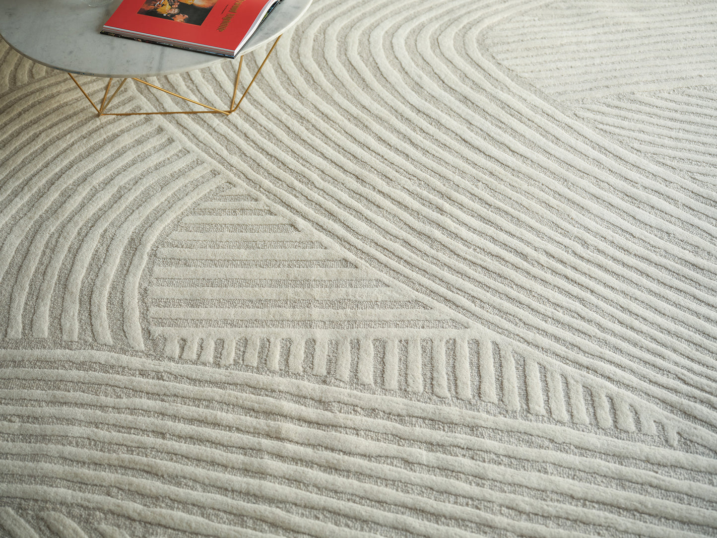 Cream rug with raised linear wave pattern; round marble table with gold legs & a red magazine on it.