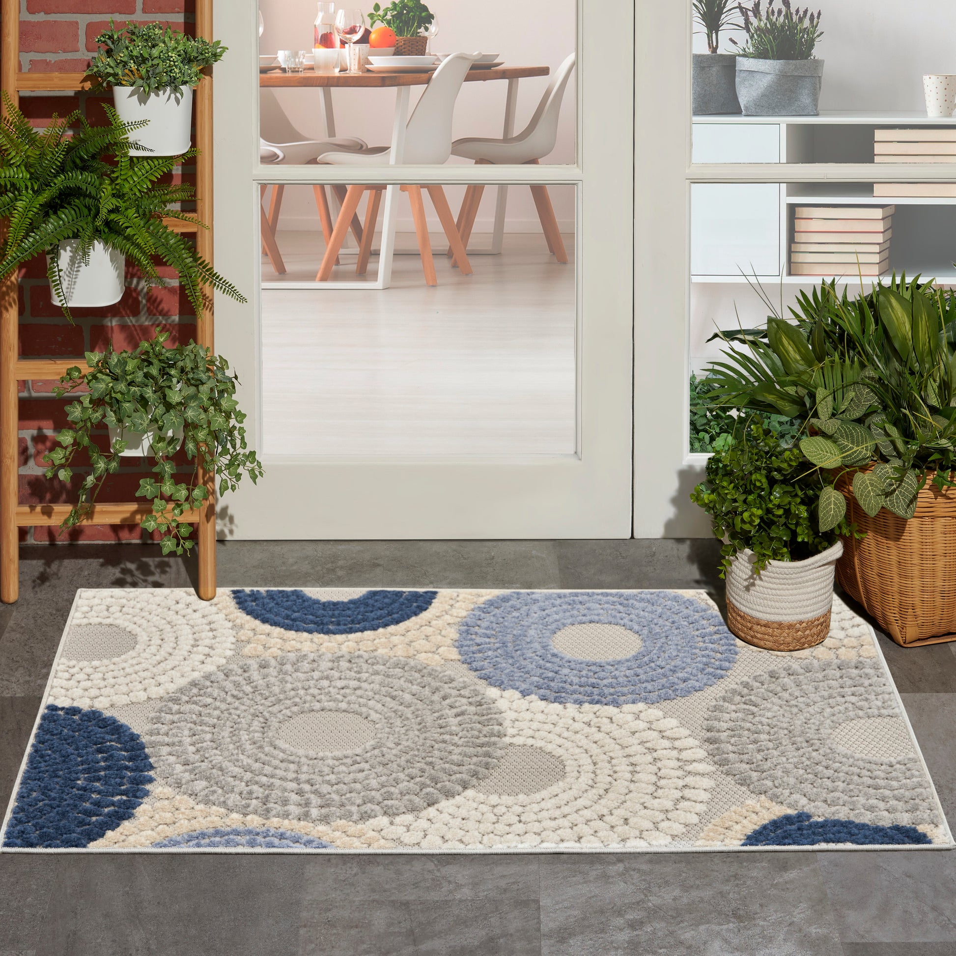 Outdoor entry rug with a circular blue, beige and gray pattern, flanked by potted plants, next to a glass door to the dining area.