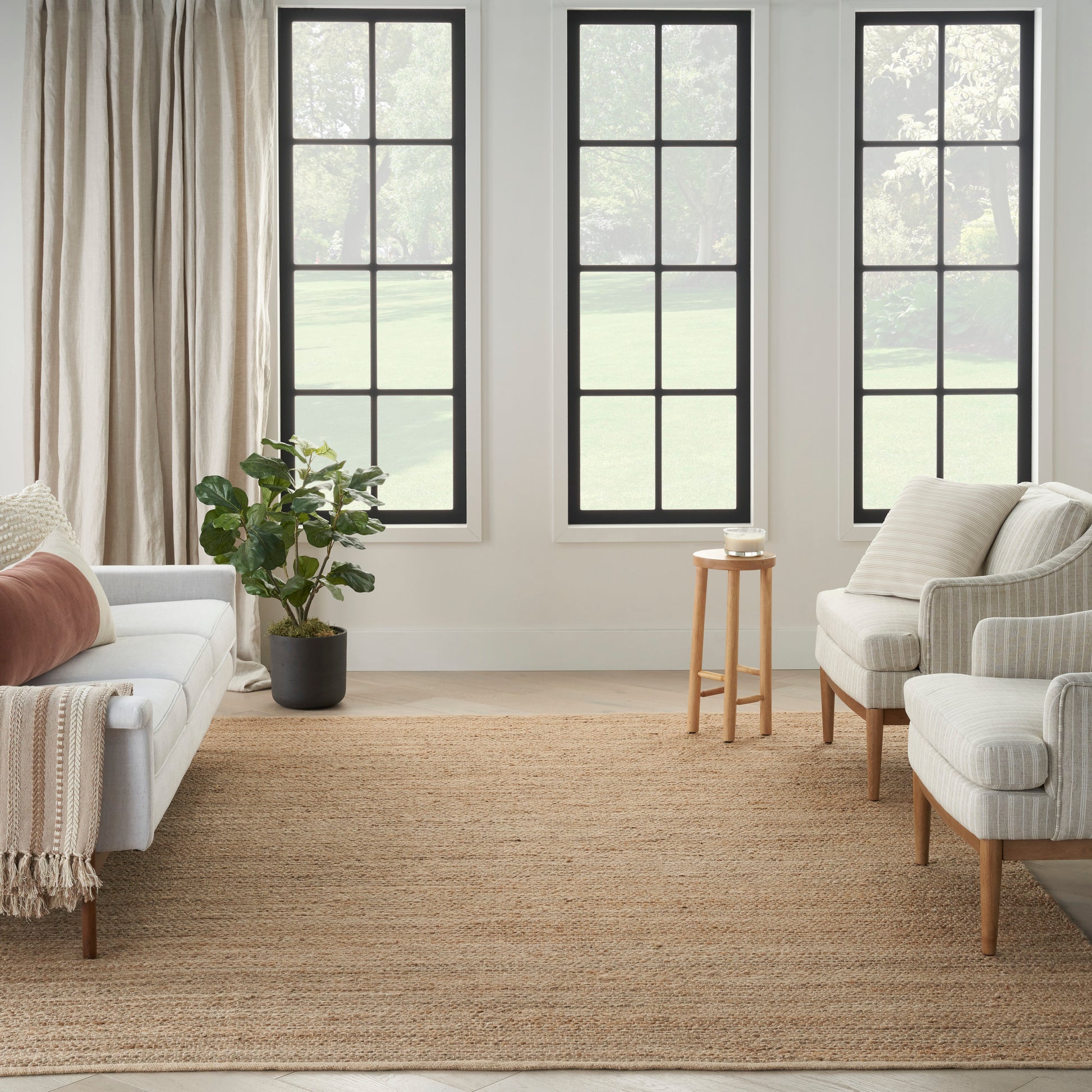 Neutral living room with black-framed windows, beige sofa, matching armchairs, jute rug, potted plant, light curtains.