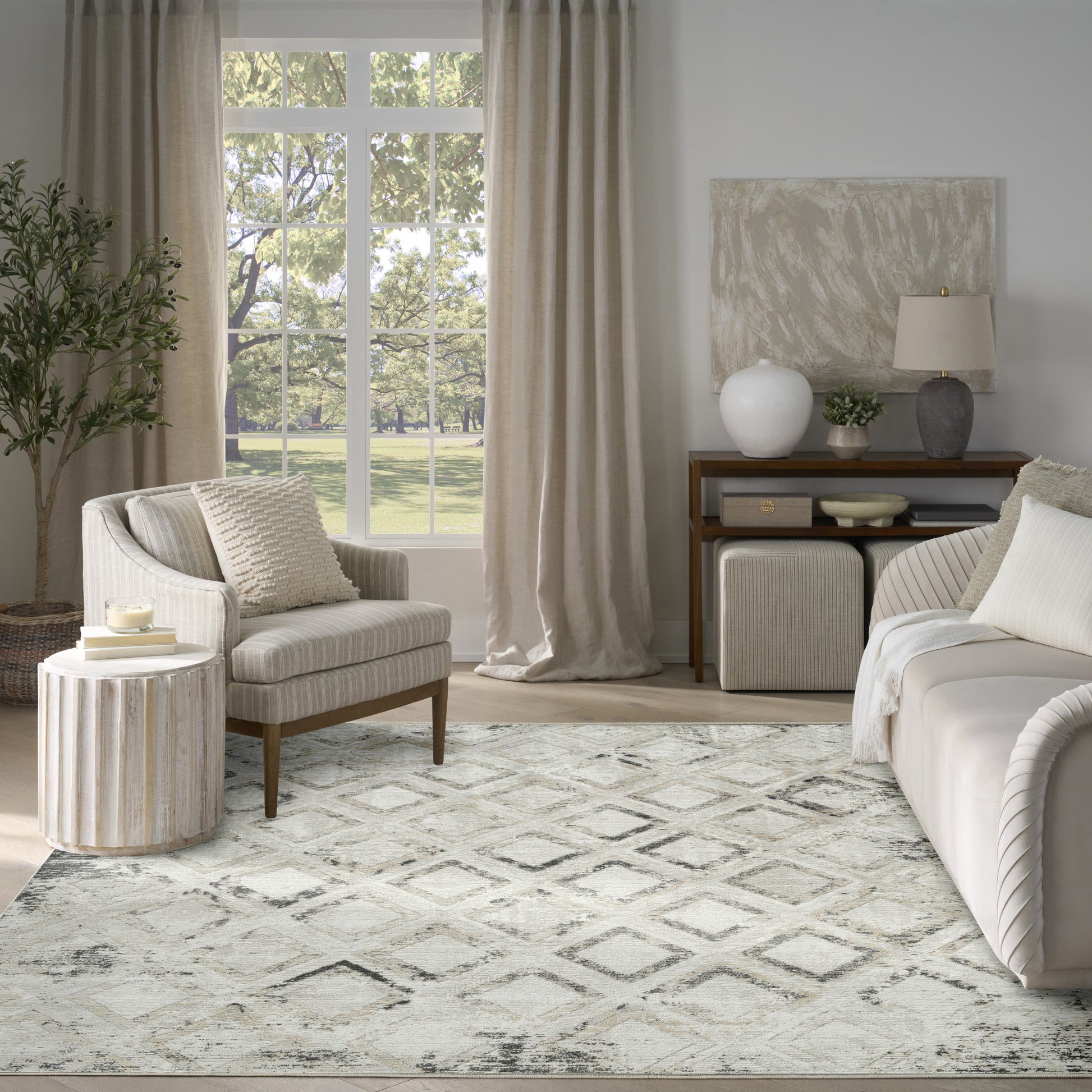 Neutral living room featuring beige sofa and armchair, diamond-pattern rug, cream curtains, wooden console and greenery.