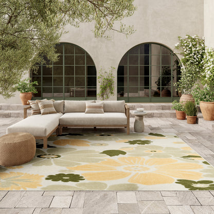 Outdoor lounge with beige modular sofa, round woven pouf, yellow-green floral rug, stone patio, arched green-framed windows.