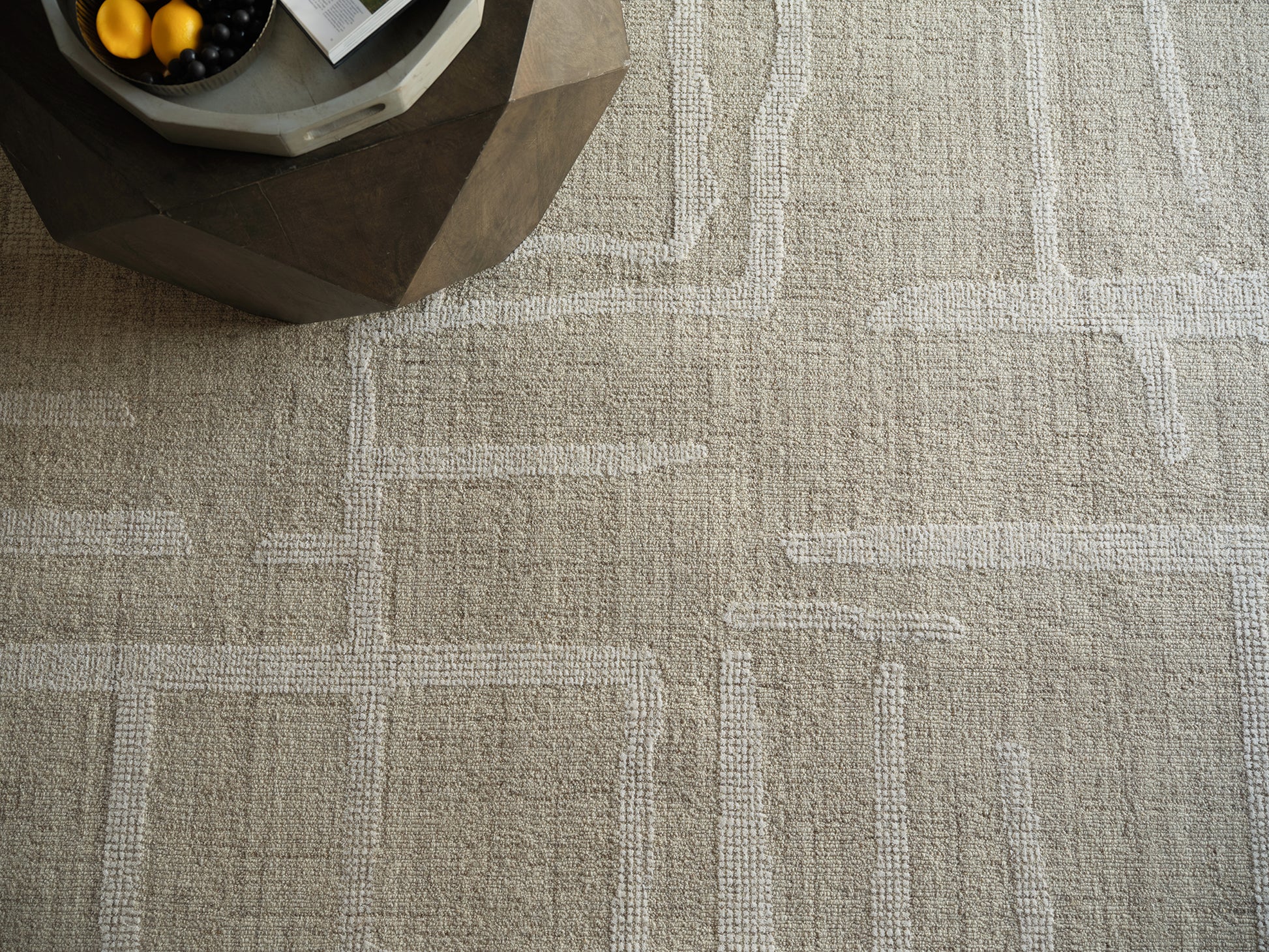 Beige geometric area rug with white rectangular borders; textured loop pile and a dark faceted coffee table in corner.