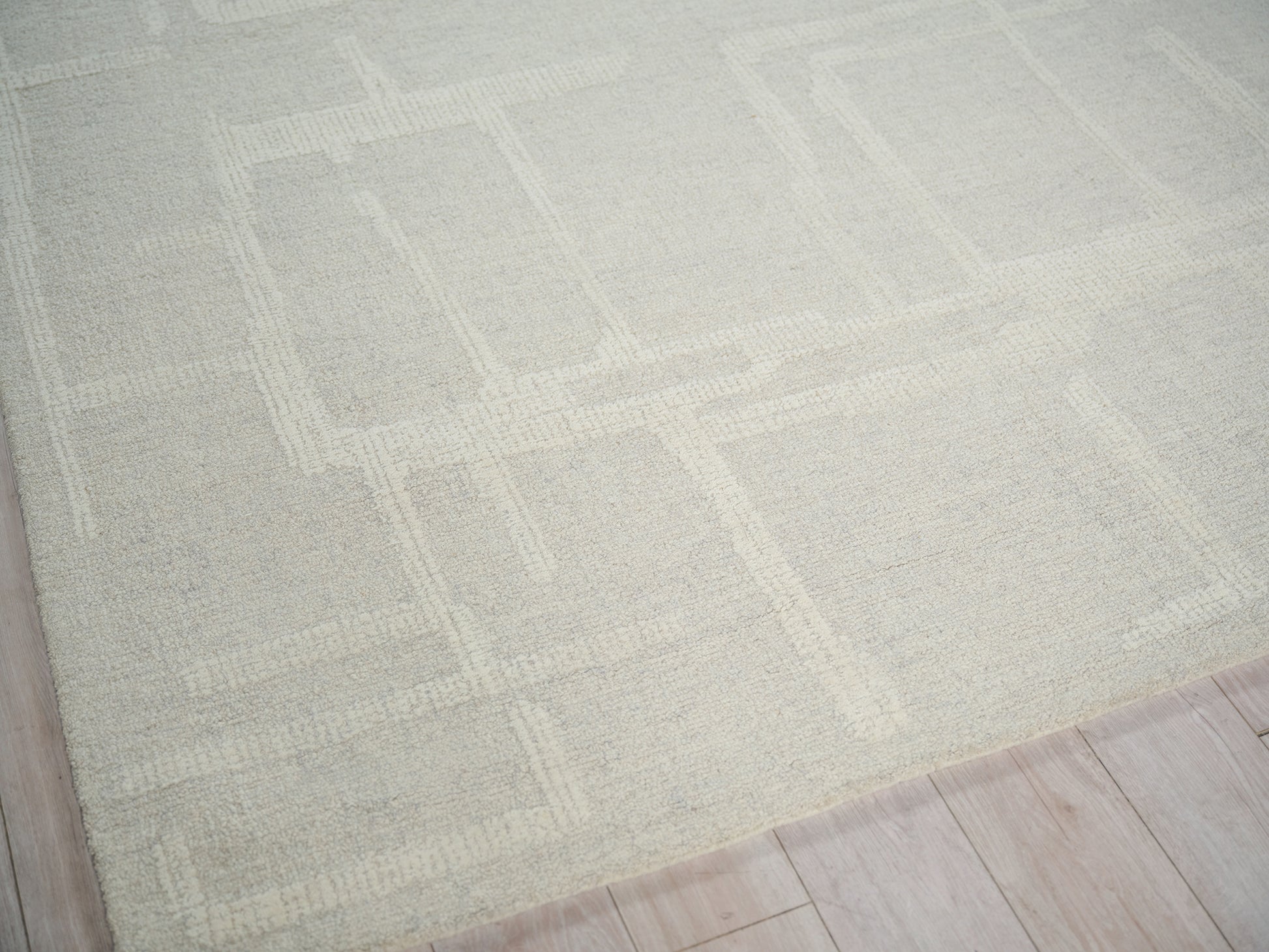 Soft beige rug with a subtle geometric square-border pattern on a light wood floor.