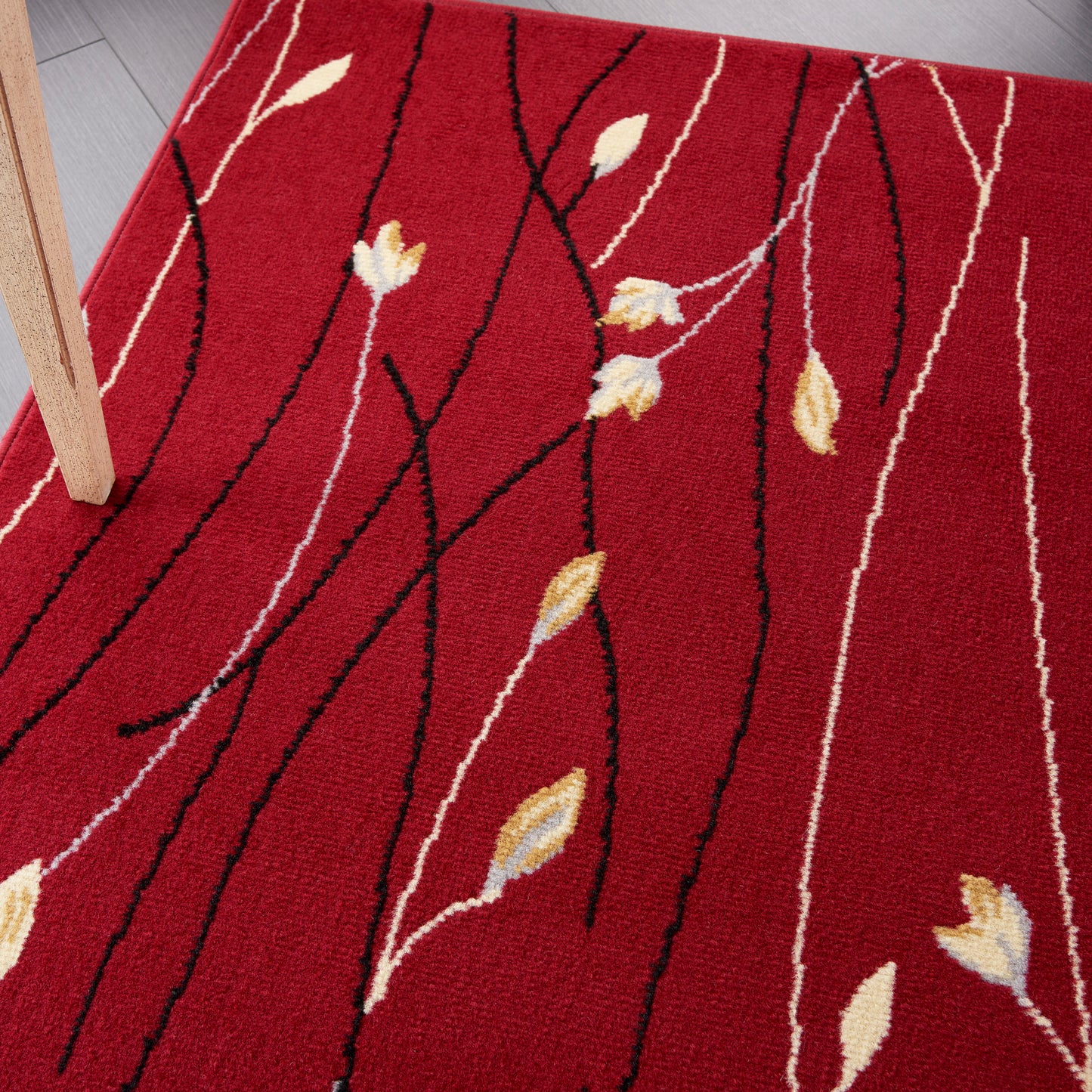 Red rug with black branch-like lines and cream-yellow leaves; modern botanical pattern under wooden furniture leg.