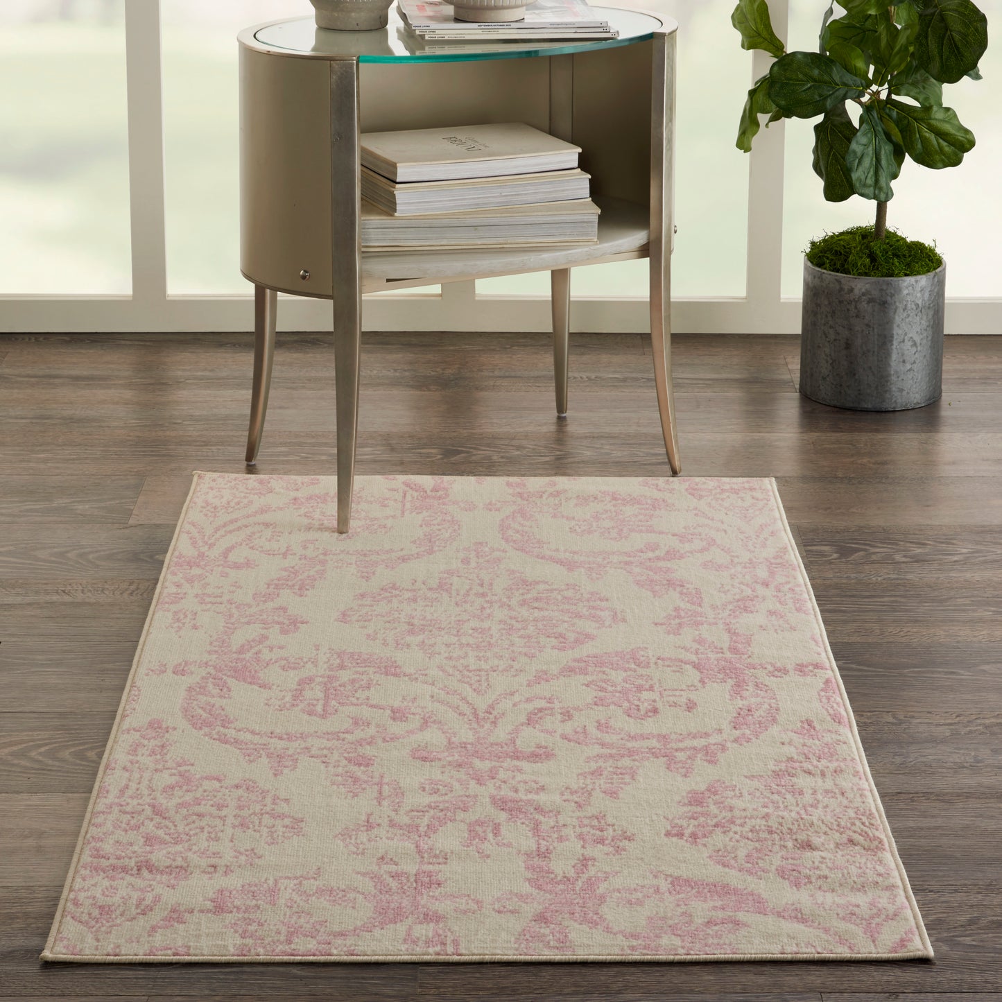 Light beige area rug with pink damask pattern on wood floor beside a round glass-top side table & potted plant