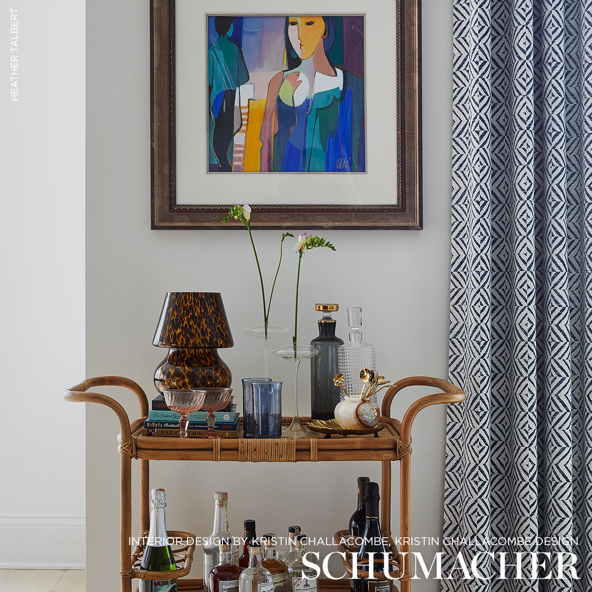 Rattan bar cart with glassware and tortoiseshell lamp, glass bottles, flower stems, abstract art, blue patterned curtains.