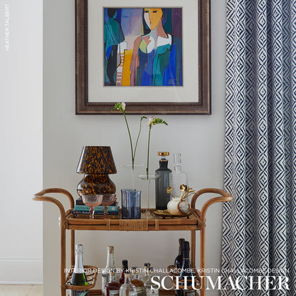 Rattan bar cart with glassware and tortoiseshell lamp, glass bottles, flower stems, abstract art, blue patterned curtains.