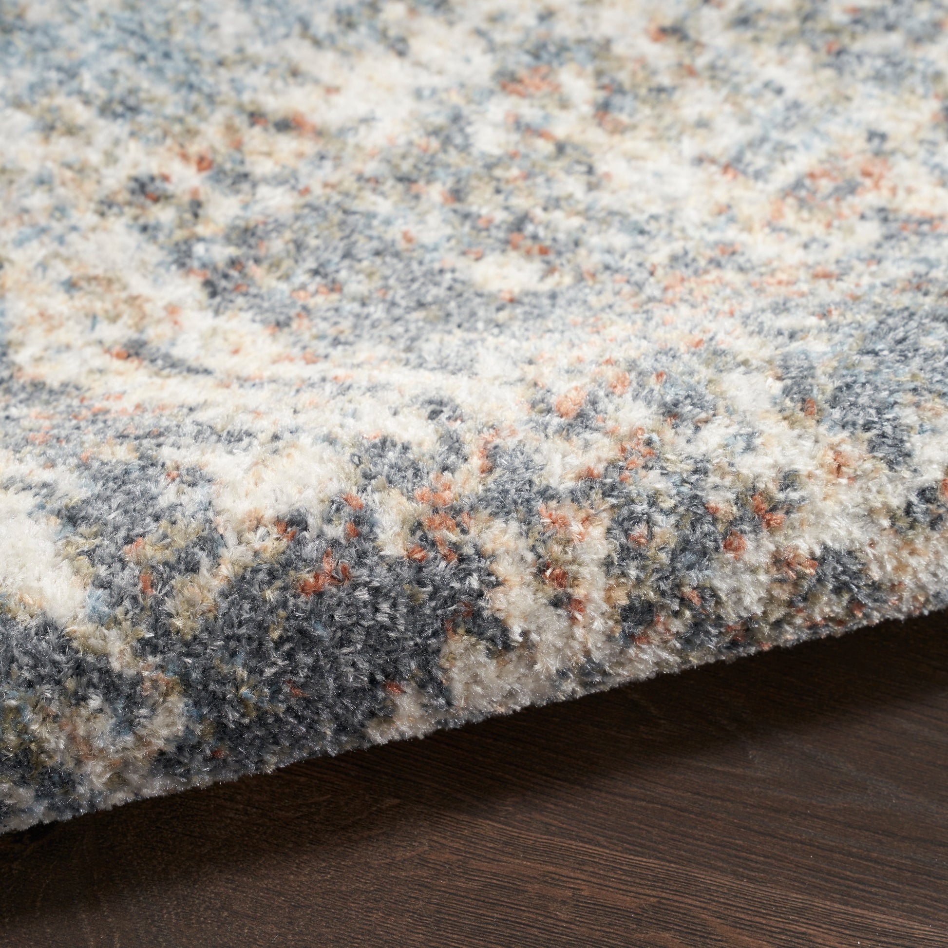 Close-up of a plush patterned rug edge on dark wood floor; blue-gray, ivory and orange flecks.
