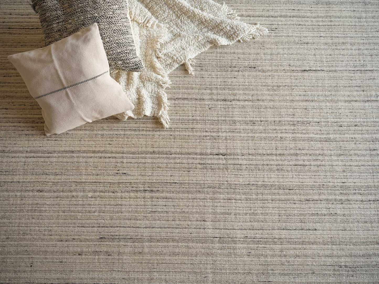 Beige cushion with dark seam and grey patterned pillow atop a cream fringed blanket on a light striped rug.