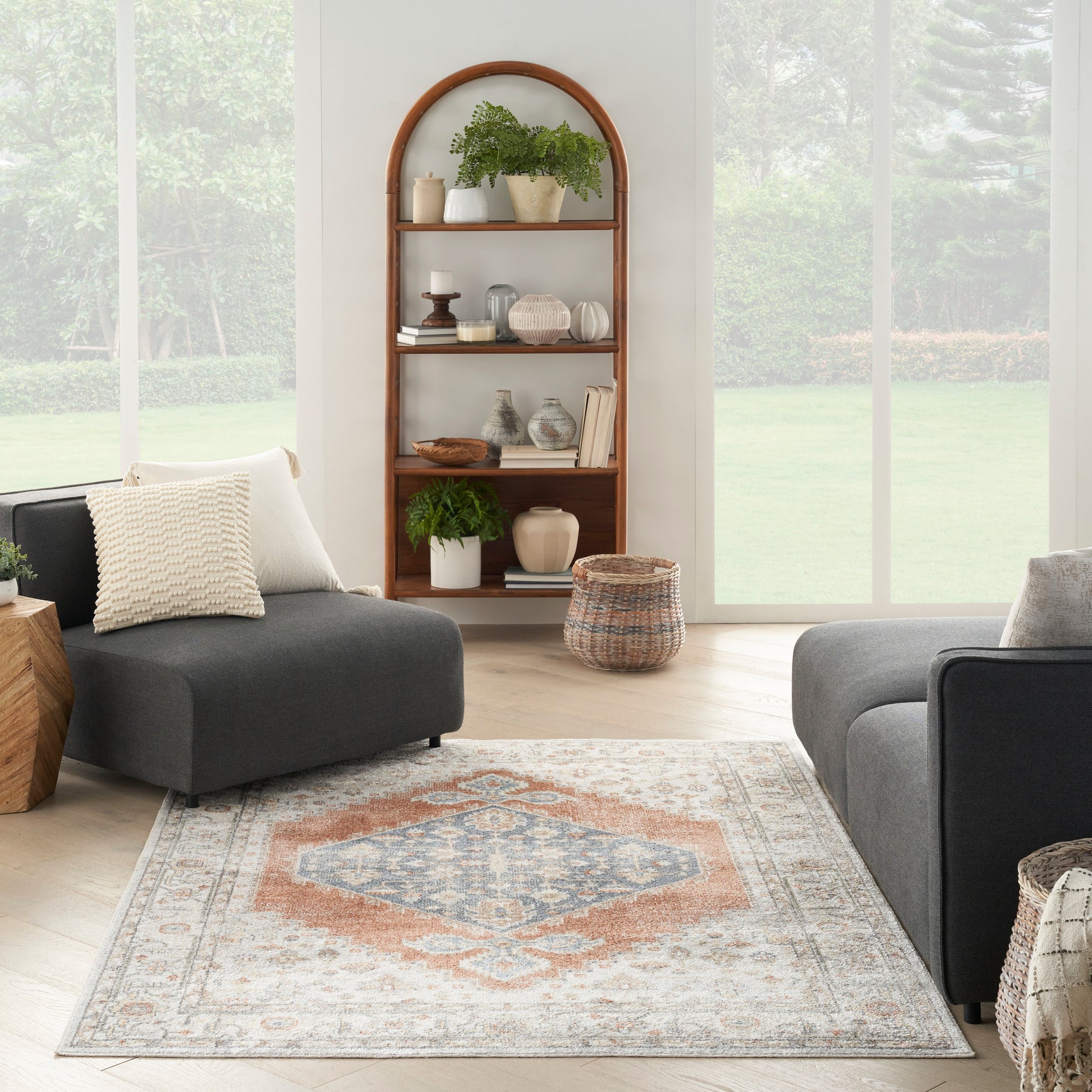 Living room with arched wooden bookshelf, gray sectional, peach-blue patterned rug, potted plants, wicker baskets.