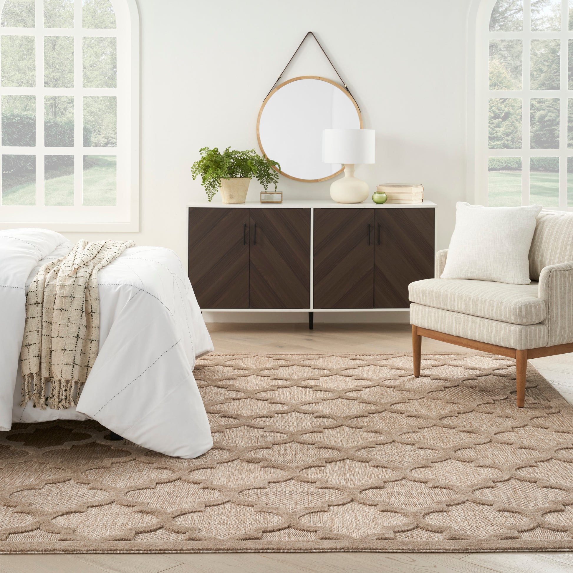 Bright bedroom with white bedding, beige knitted throw, dark chevron cabinet, round wood mirror, plant, lamp, beige rug.