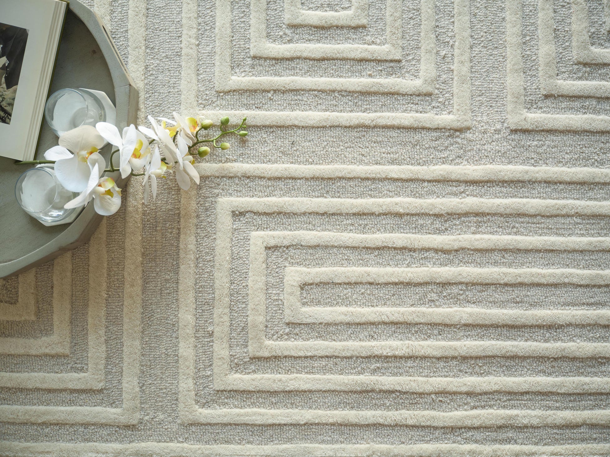 Beige textured rug with raised concentric square pattern; round tray on left with a book, glass items & white orchid blossoms.