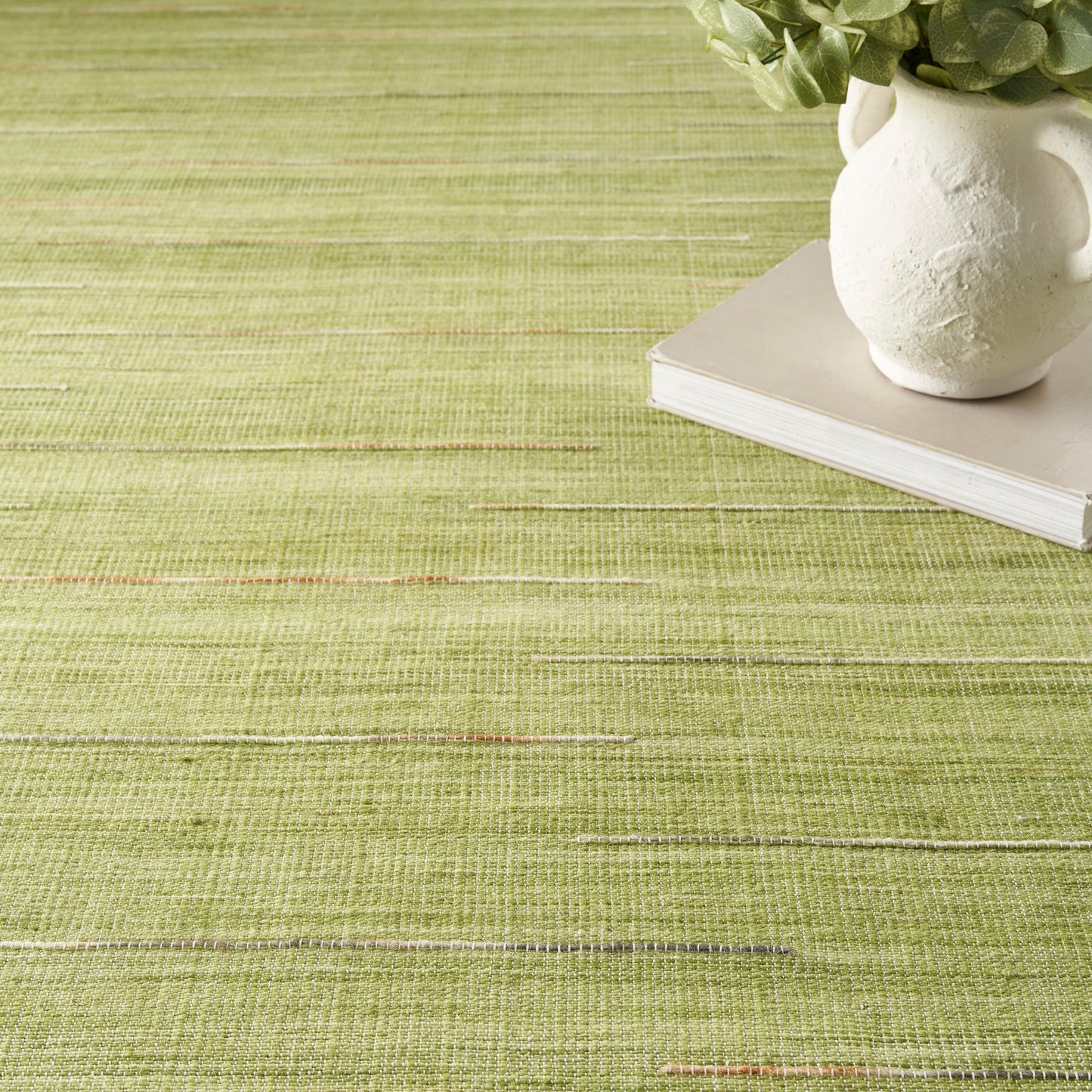Olive-green woven rug with a white ceramic vase & plant atop a book.