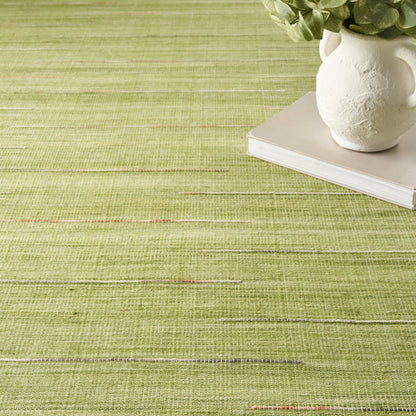 Olive-green woven rug with a white ceramic vase & plant atop a book.