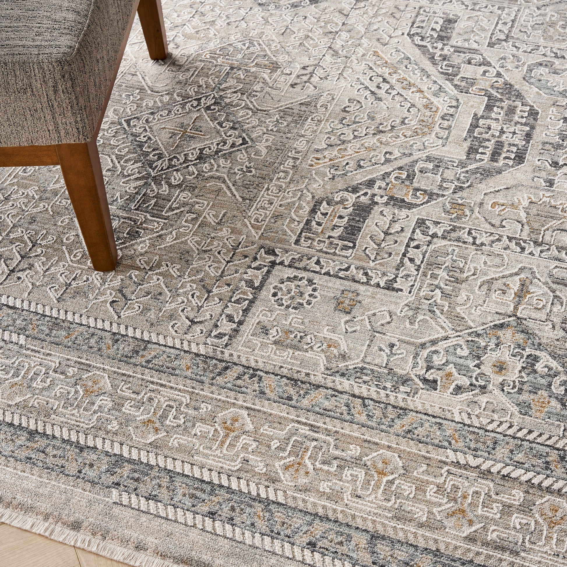 Neutral geometric patterned area rug in beige-gray tones with ornate borders; wooden chair leg visible.