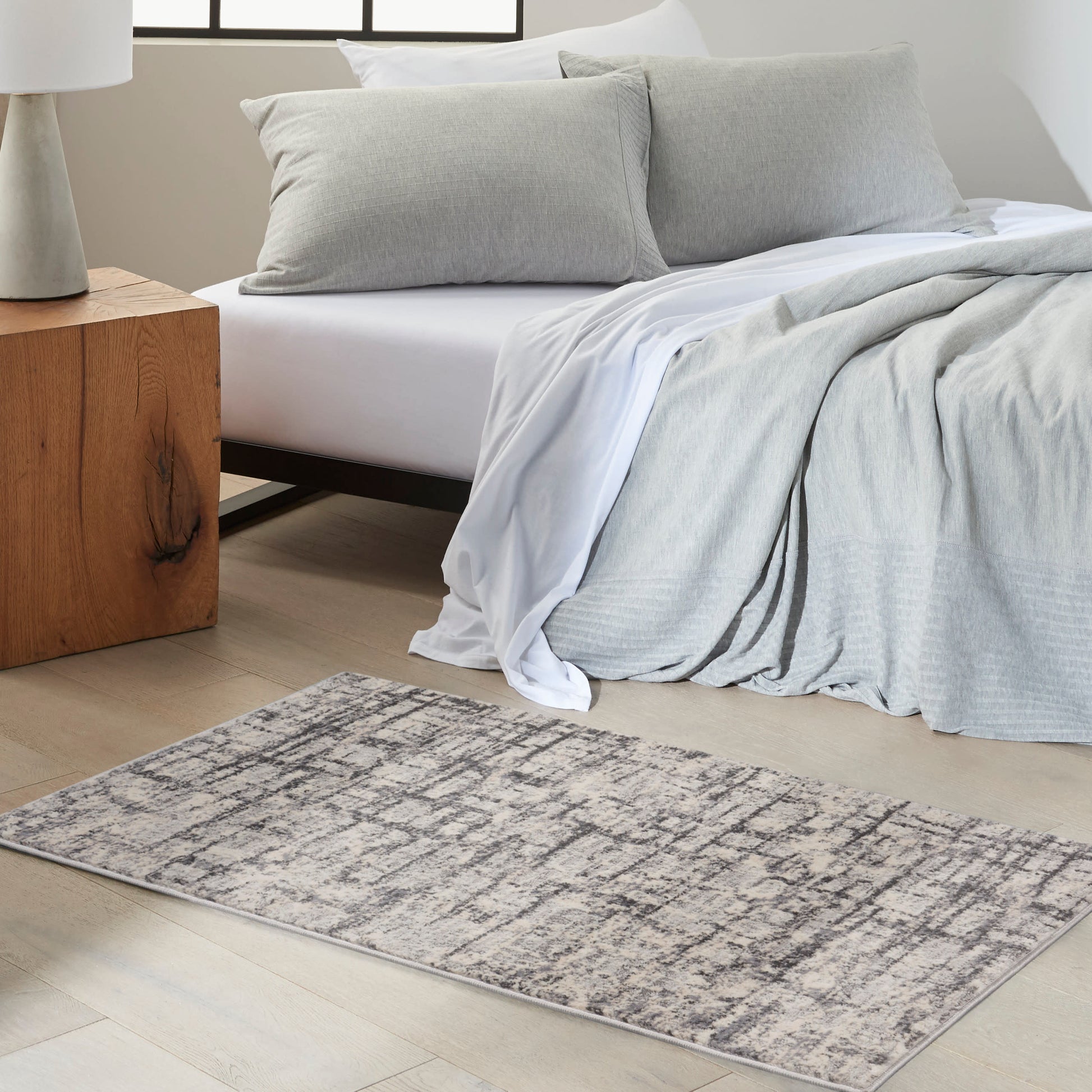 Gray bedding with white sheets on a bed, modern gray rug with abstract pattern; wooden block nightstand with lamp.