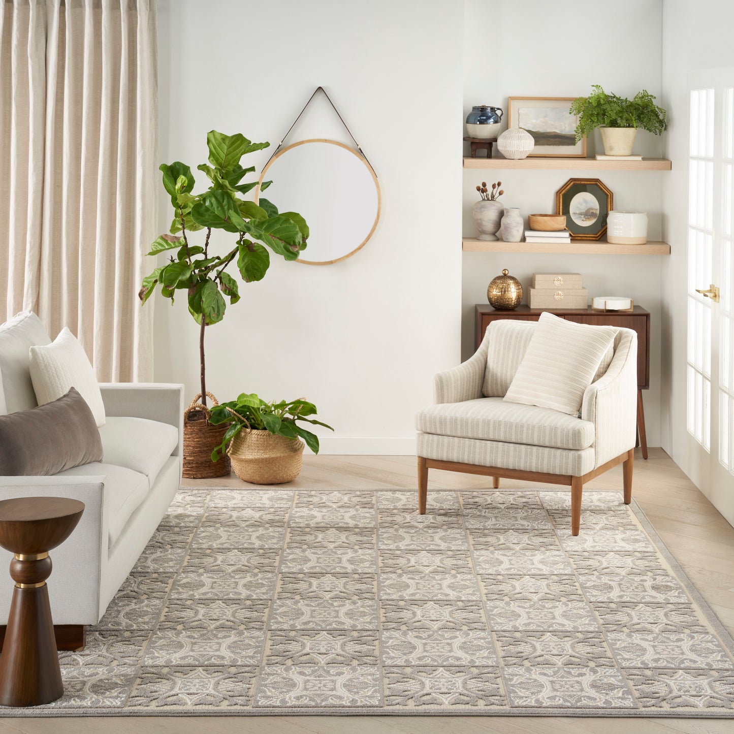 Neutral living room with light sofa, striped armchair, gray patterned rug, large potted plant, round wall hoop, and shelves.