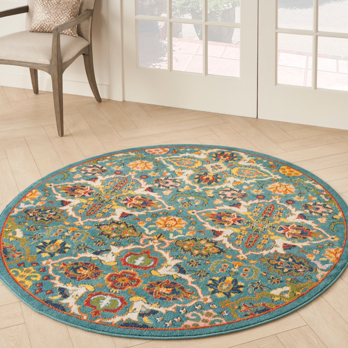 Round turquoise rug with intricate multicolor floral pattern; beige chair and glass doors in a sunlit wood floor room.