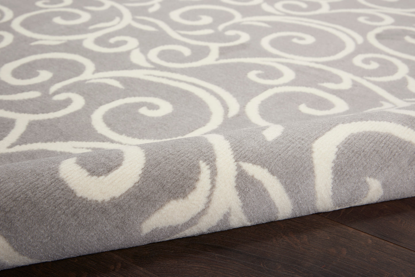 Soft gray area rug with white swirling scroll pattern, plush texture, edge folded on dark hardwood floor.