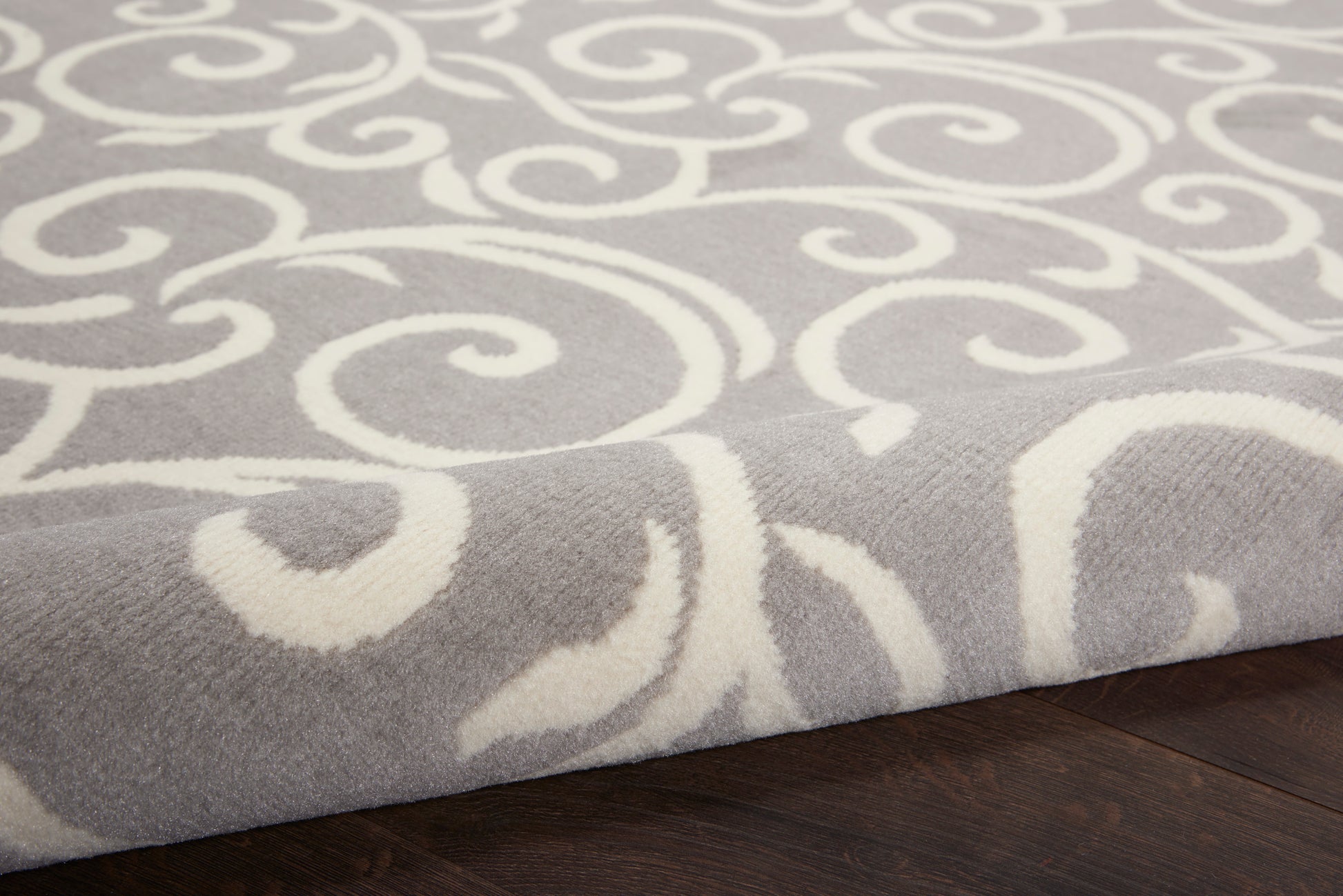 Soft gray area rug with white swirling scroll pattern, plush texture, edge folded on dark hardwood floor.