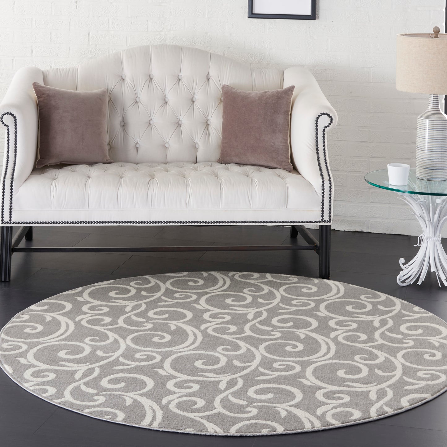 Cream tufted loveseat with dark legs and nailhead trim, mauve throw pillows, round gray swirl rug, glass side table.