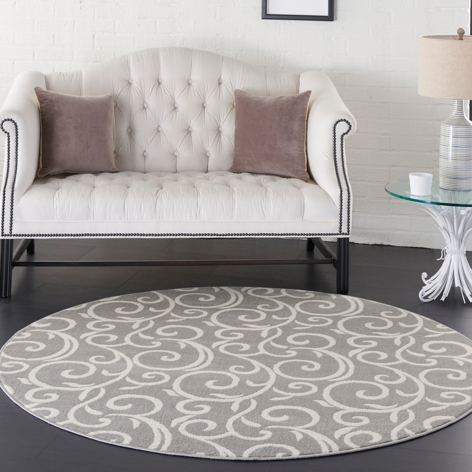 Cream tufted loveseat with dark legs and nailhead trim, mauve throw pillows, round gray swirl rug, glass side table.