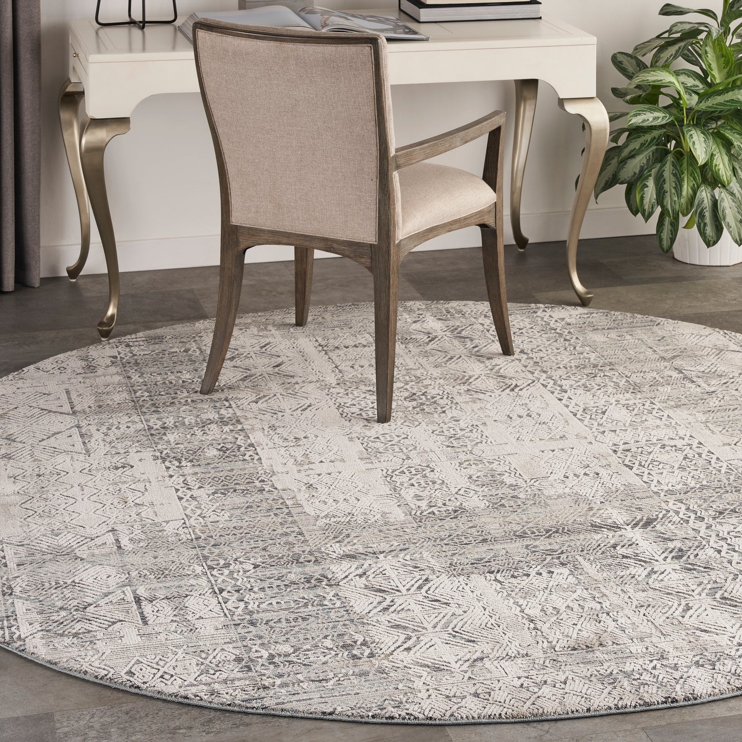 Round taupe-gray geometric rug under beige upholstered chair and cream desk with curved gold legs.