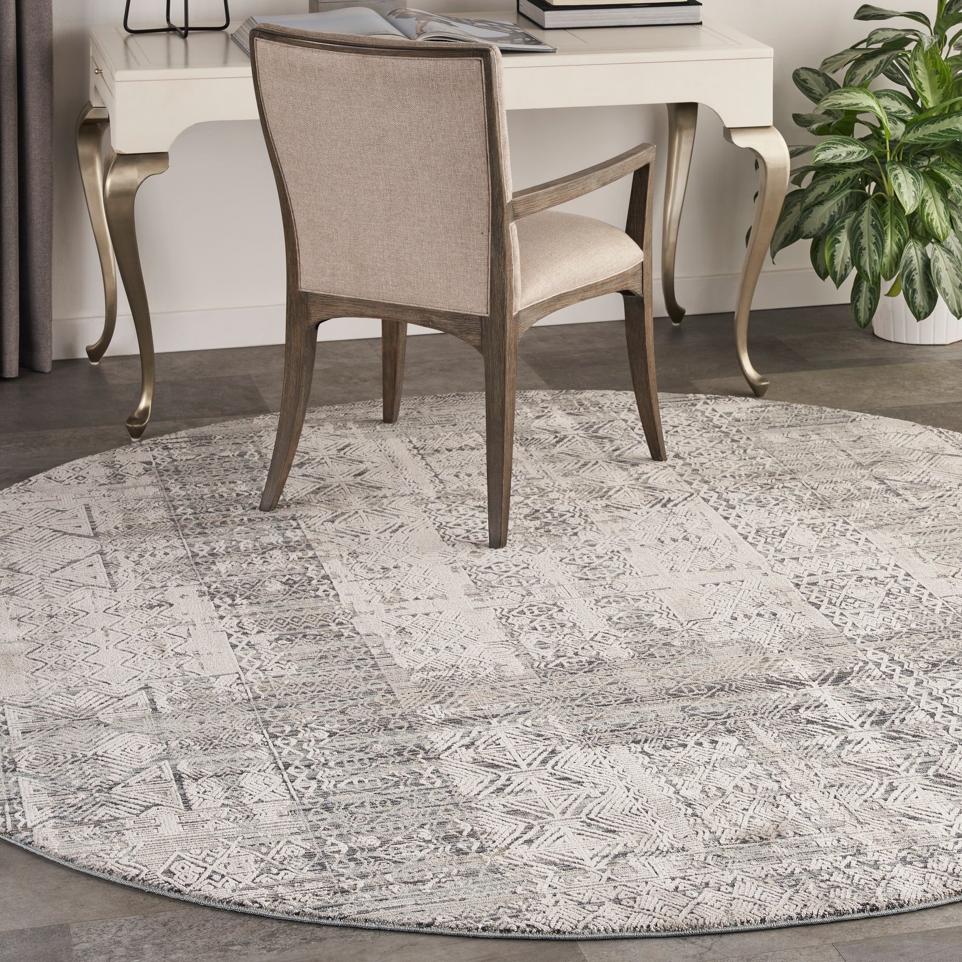 Round taupe-gray geometric rug under beige upholstered chair and cream desk with curved gold legs.