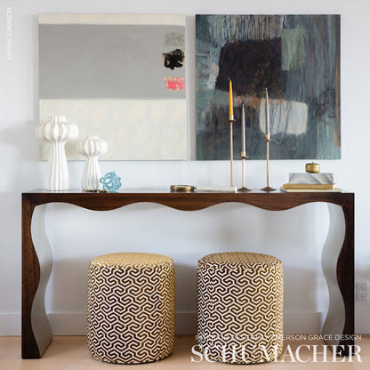Dark wood console with curved legs, two beige-black patterned poufs, abstract wall art, white ceramic lamps, candles, books.