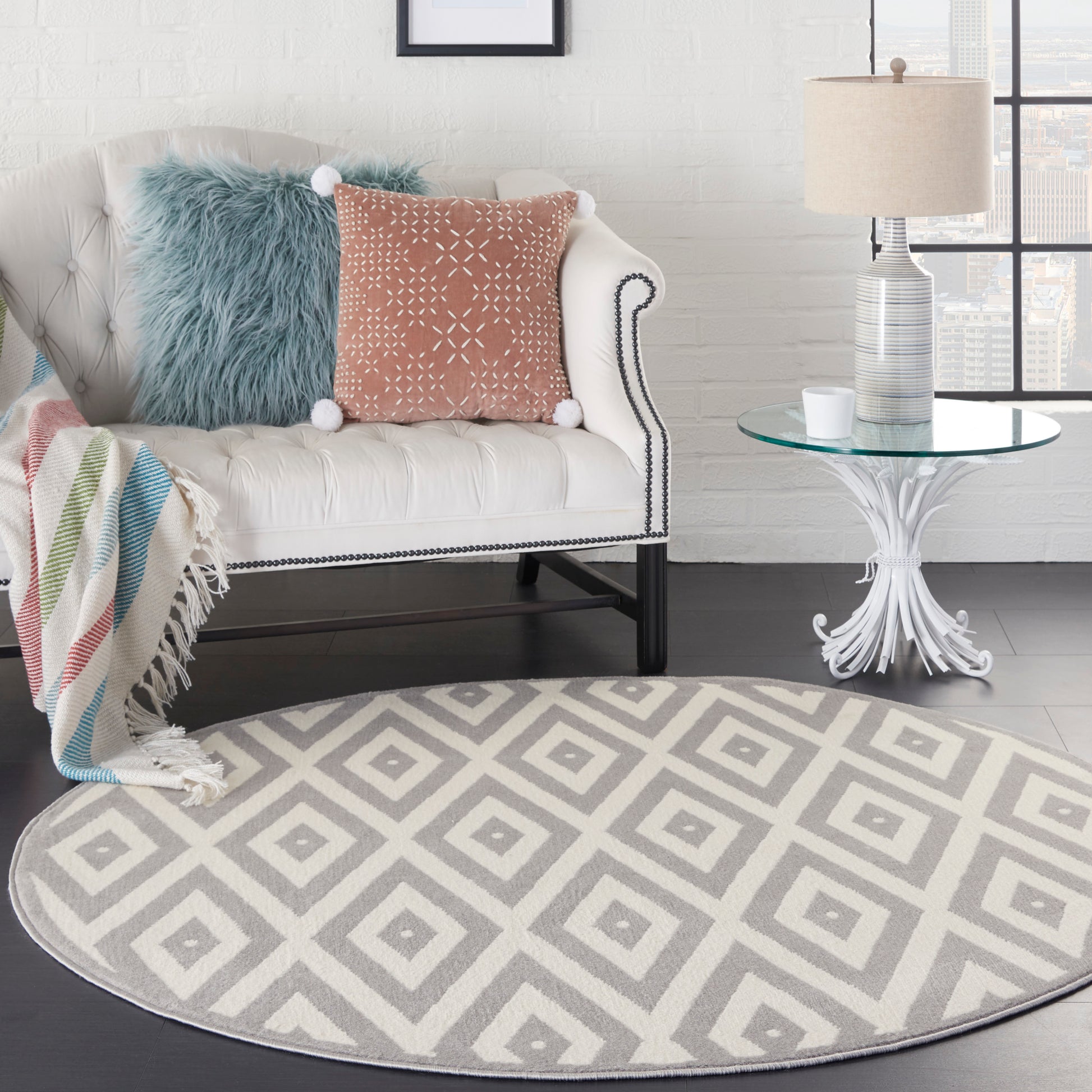 Round gray geometric rug under white tufted sofa with nailhead trim, blue faux-fur and pink pom-pom pillows, glass side table