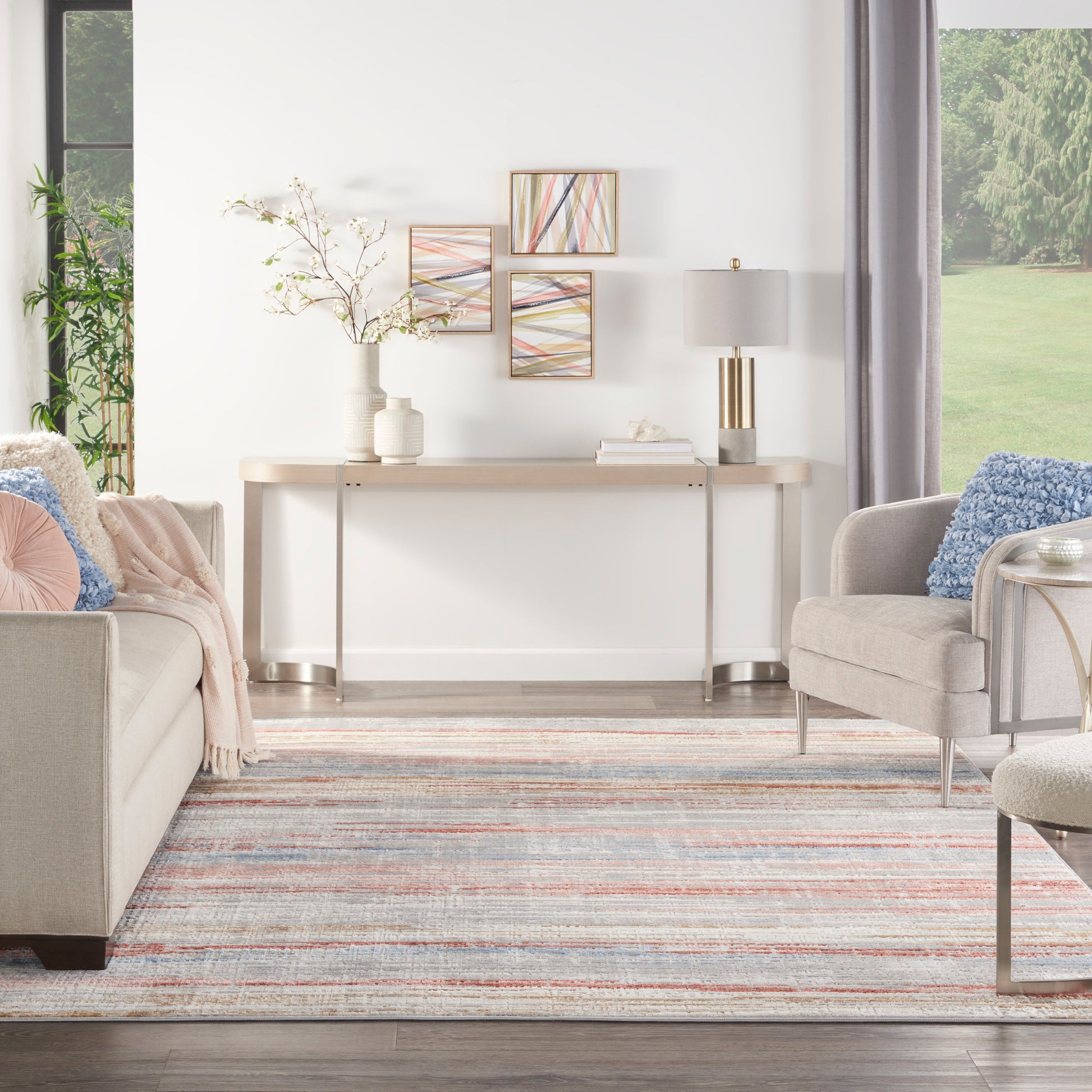 Bright living room with light wood console, beige sofa, gray armchair, pastel striped rug, and abstract art above console.