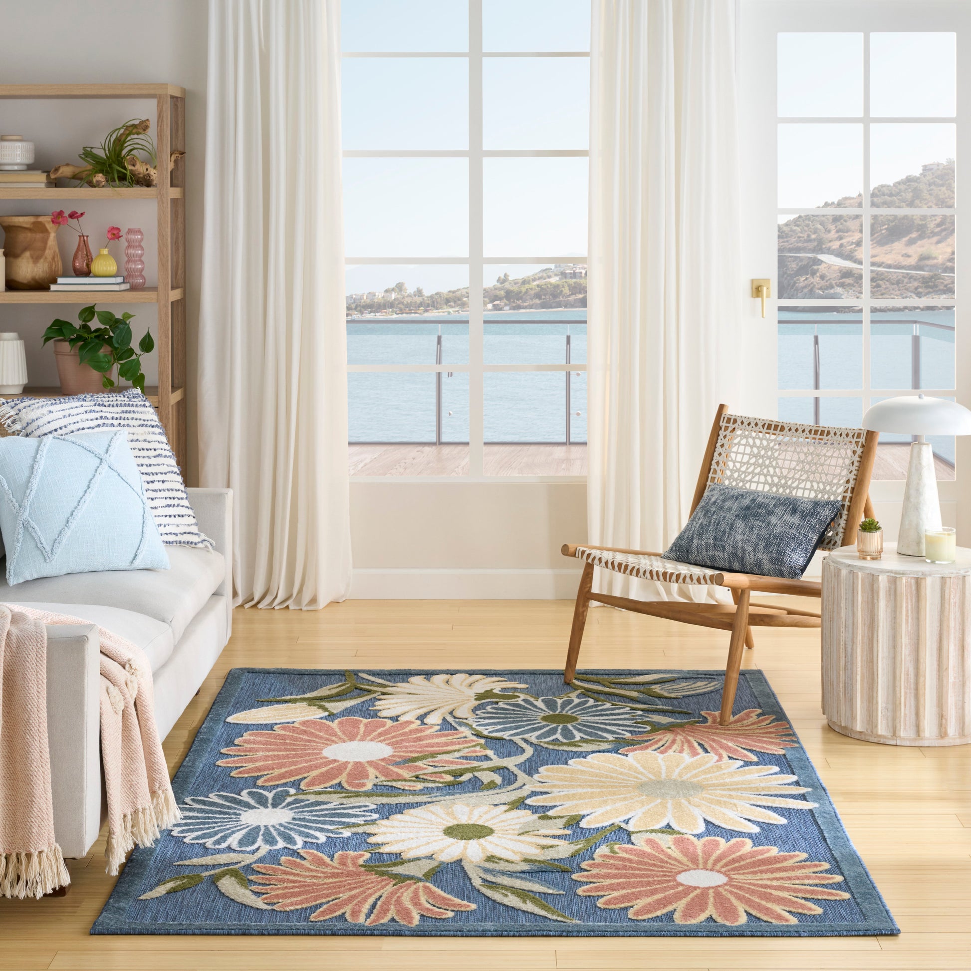 Outdoor coastal patio scene with a blue floral area rug, cream sofa, woven chair, light wood deck, and sea view.