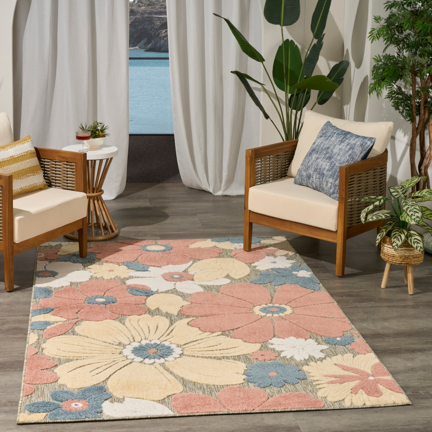 Pastel floral area rug with pink, cream, blue and yellow flowers, paired with wooden-framed wicker chairs and indoor plants.