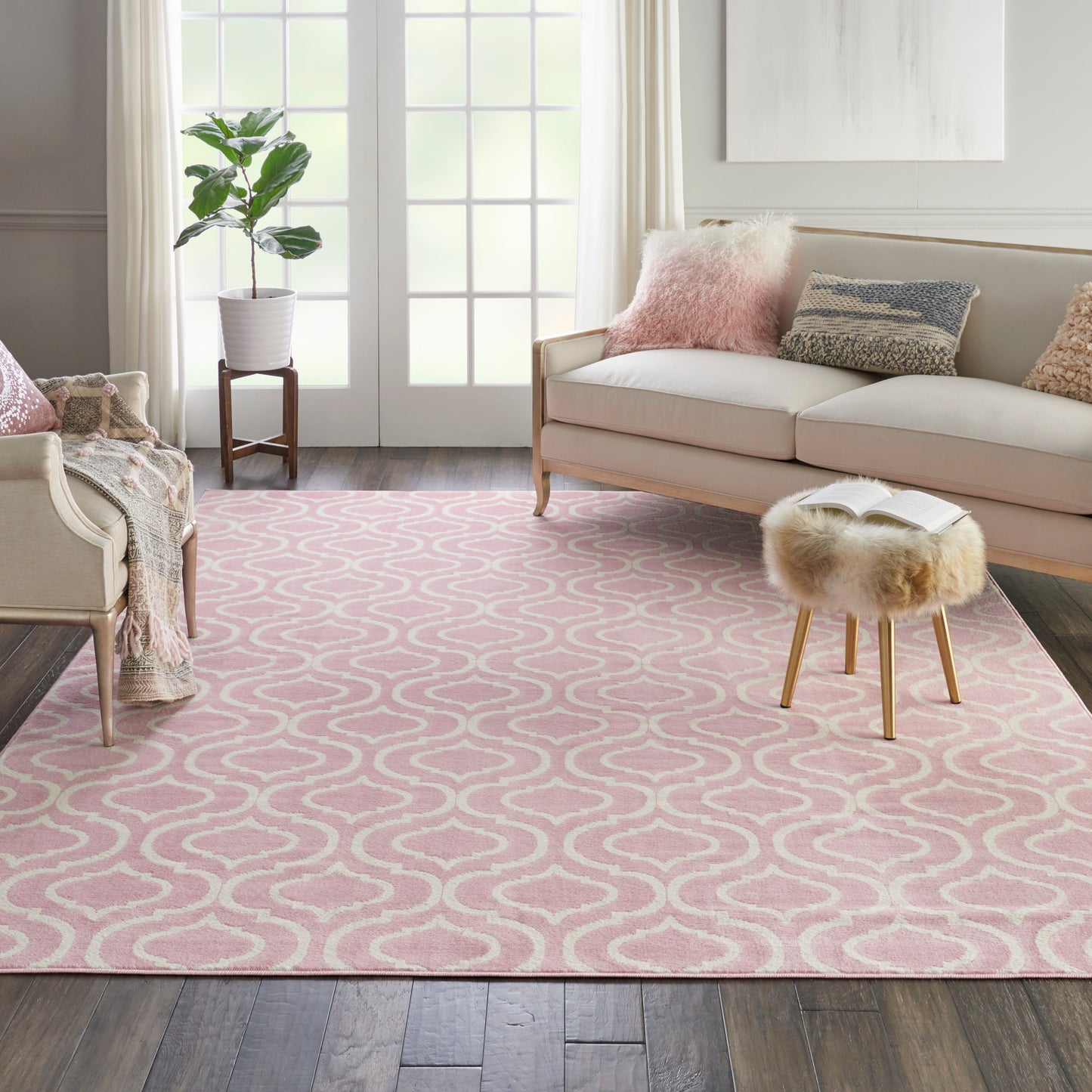 Pink area rug with white wavy lattice pattern in a bright living room with beige sofa, fur ottoman, and plants.