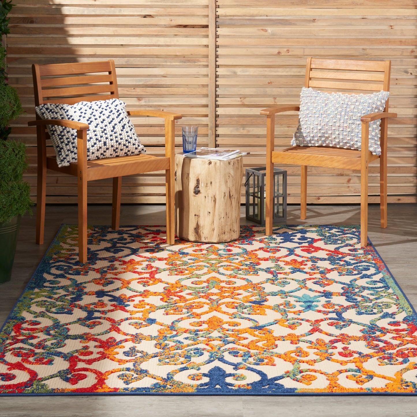 Outdoor wooden chairs with blue pillows flank a tree-stump table on a vibrant multicolor patterned rug beside a wooden fence.