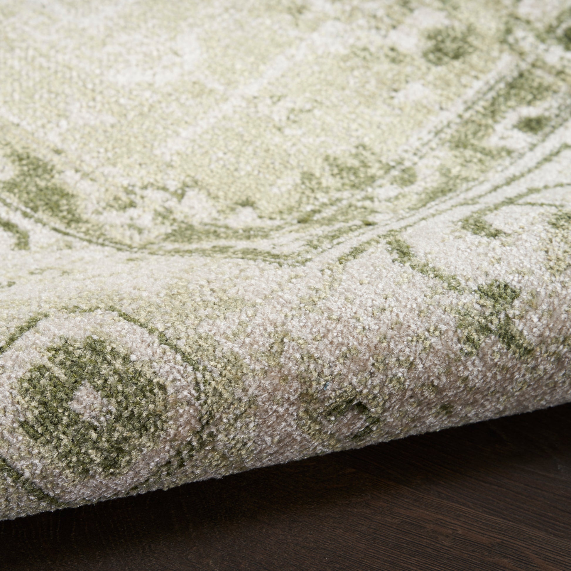 Close-up of a plush green & ivory patterned rug with a rolled edge on dark wood floor.