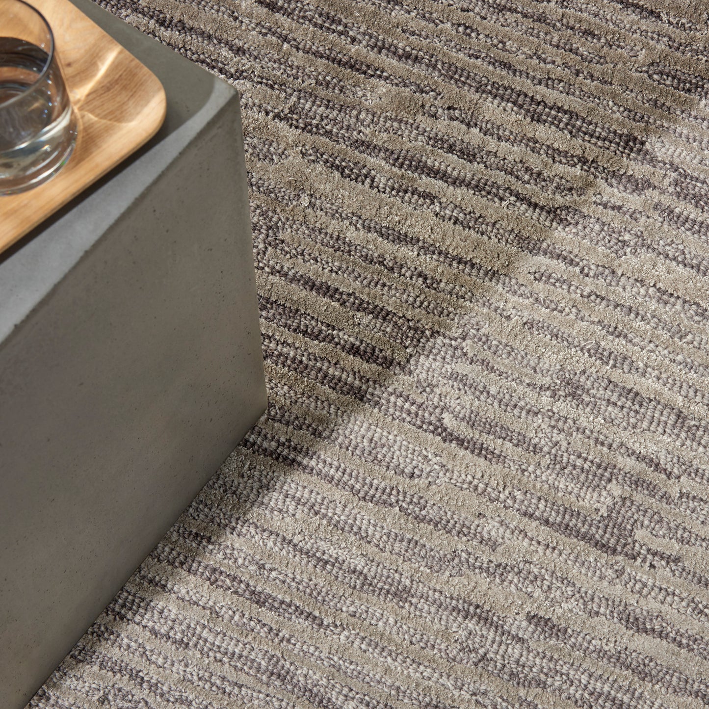 Gray concrete block table with a light wood tray and glass, set on a textured striped gray rug.