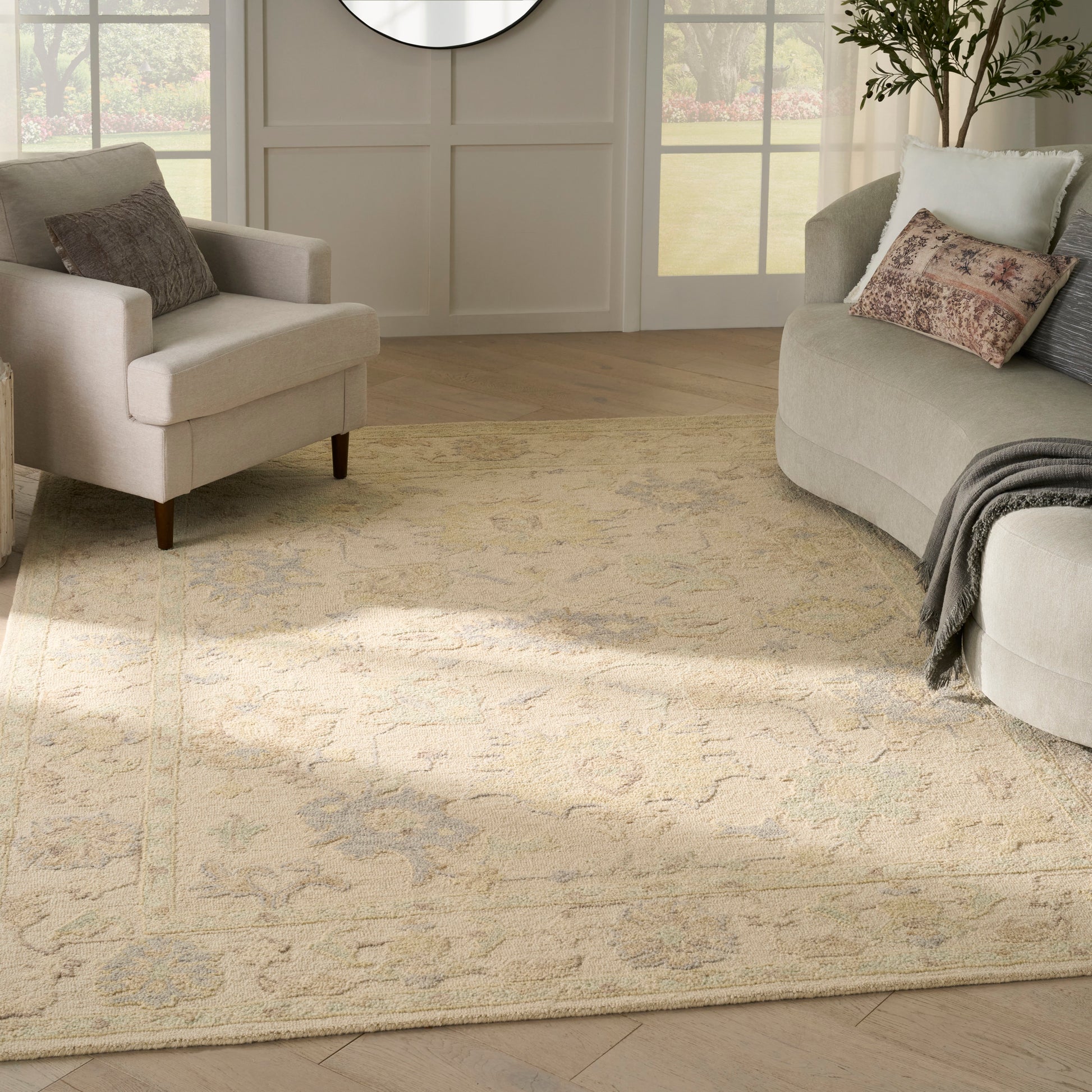 Neutral living room with beige patterned area rug, light gray curved sofa, beige armchair, round wall mirror, pillows.