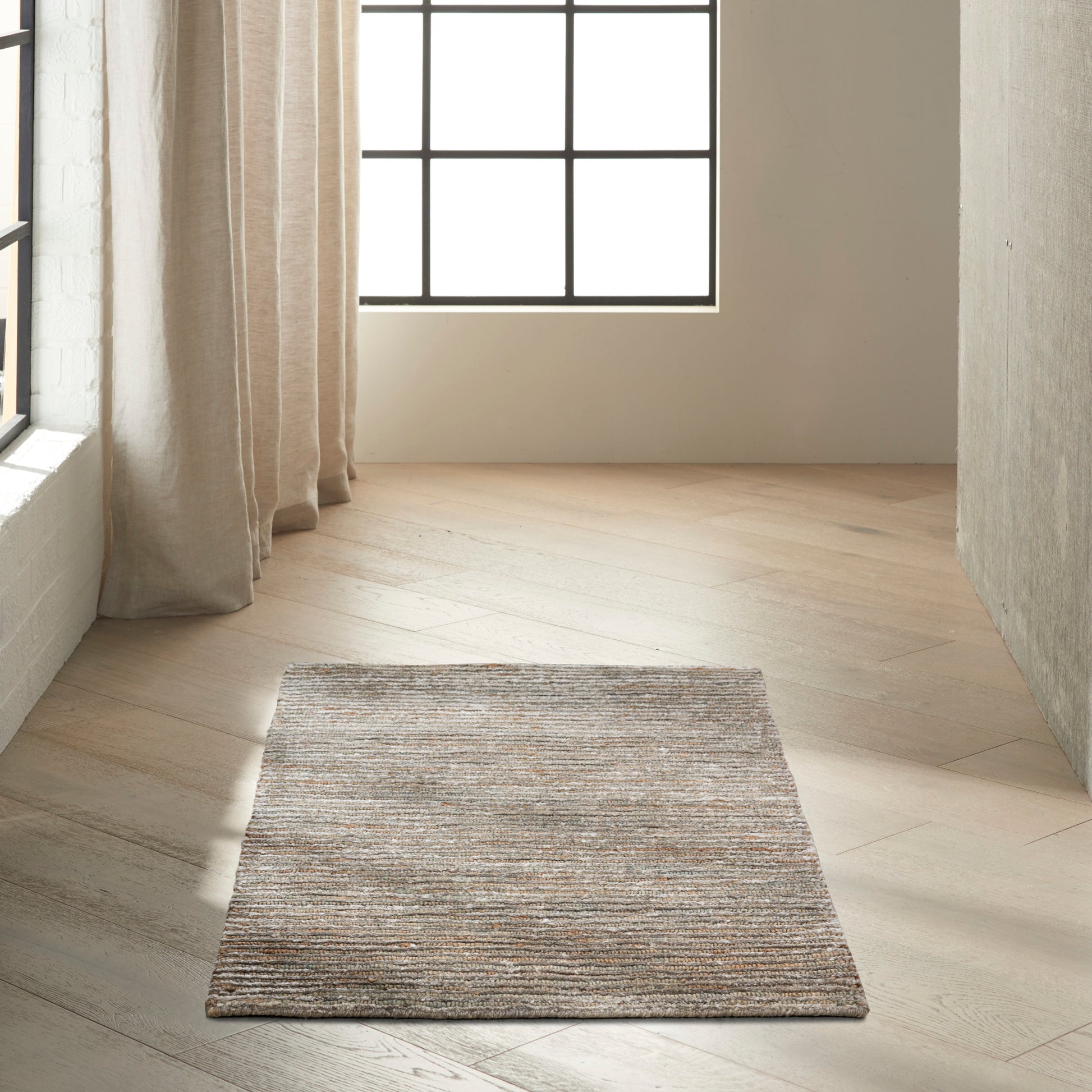 Neutral woven rug in beige-gray tones placed on a light wood floor beside a large grid window with sheer curtains.