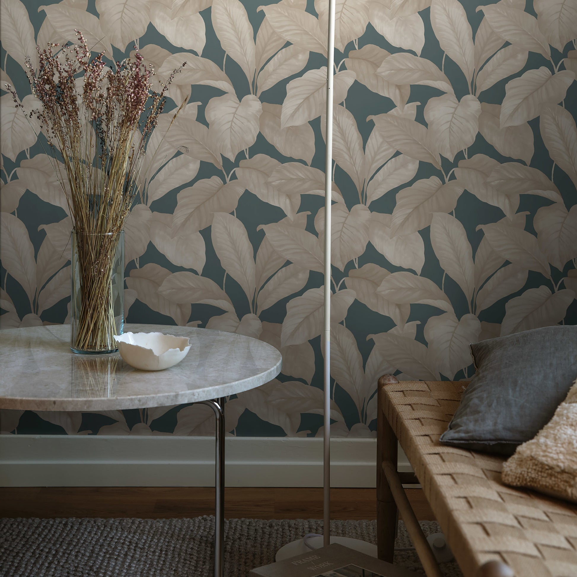 Round marble-top side table with metal legs, vase of dried stems, teal botanical wallpaper, woven sofa.