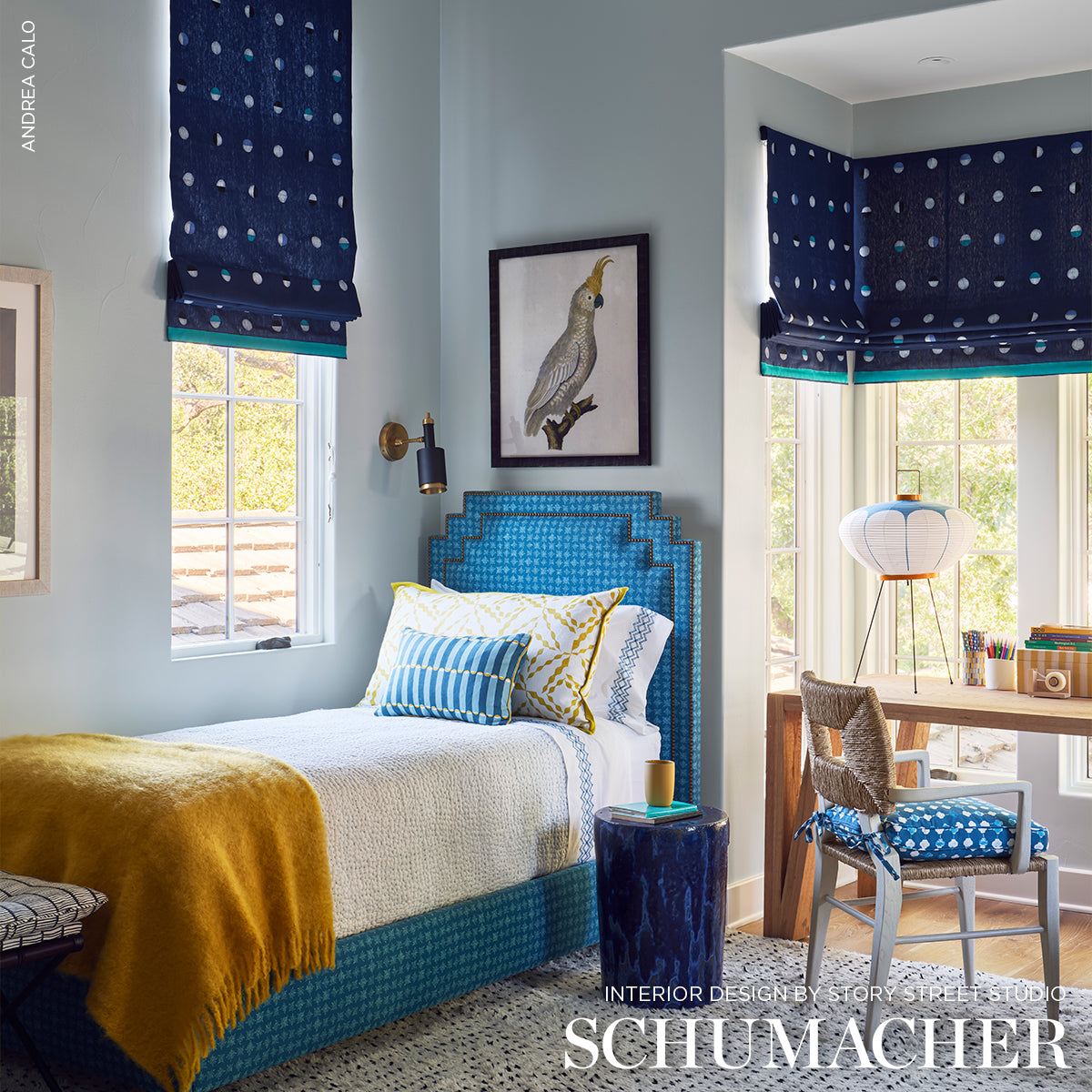 Indigo moons woven wallpaper on a bright mid-century bedroom wall; blue turquoise upholstered bed with geometric headboard, yellow throw, blue dotted roman blinds, desk by the window.