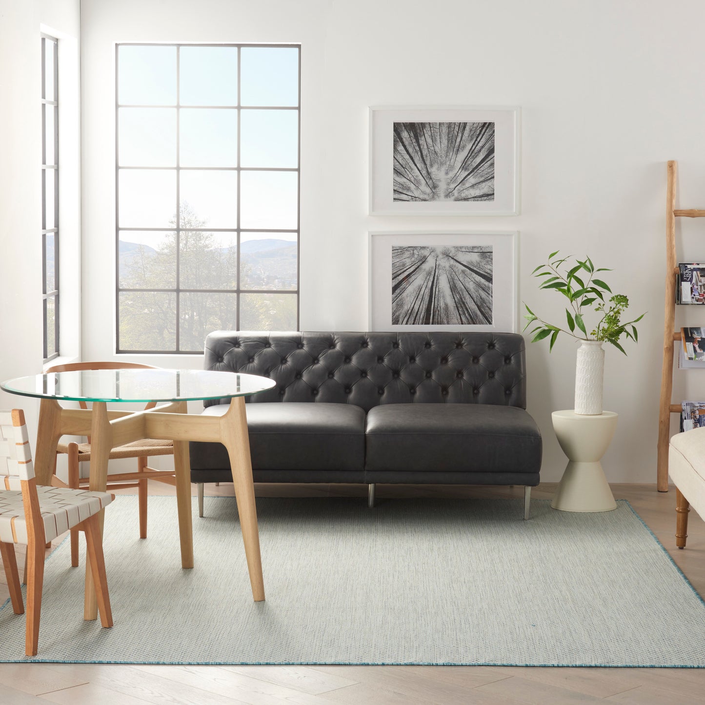 Contemporary living room with black tufted leather sofa, glass table, light wood chairs, grid window, plant & black-and-white art.