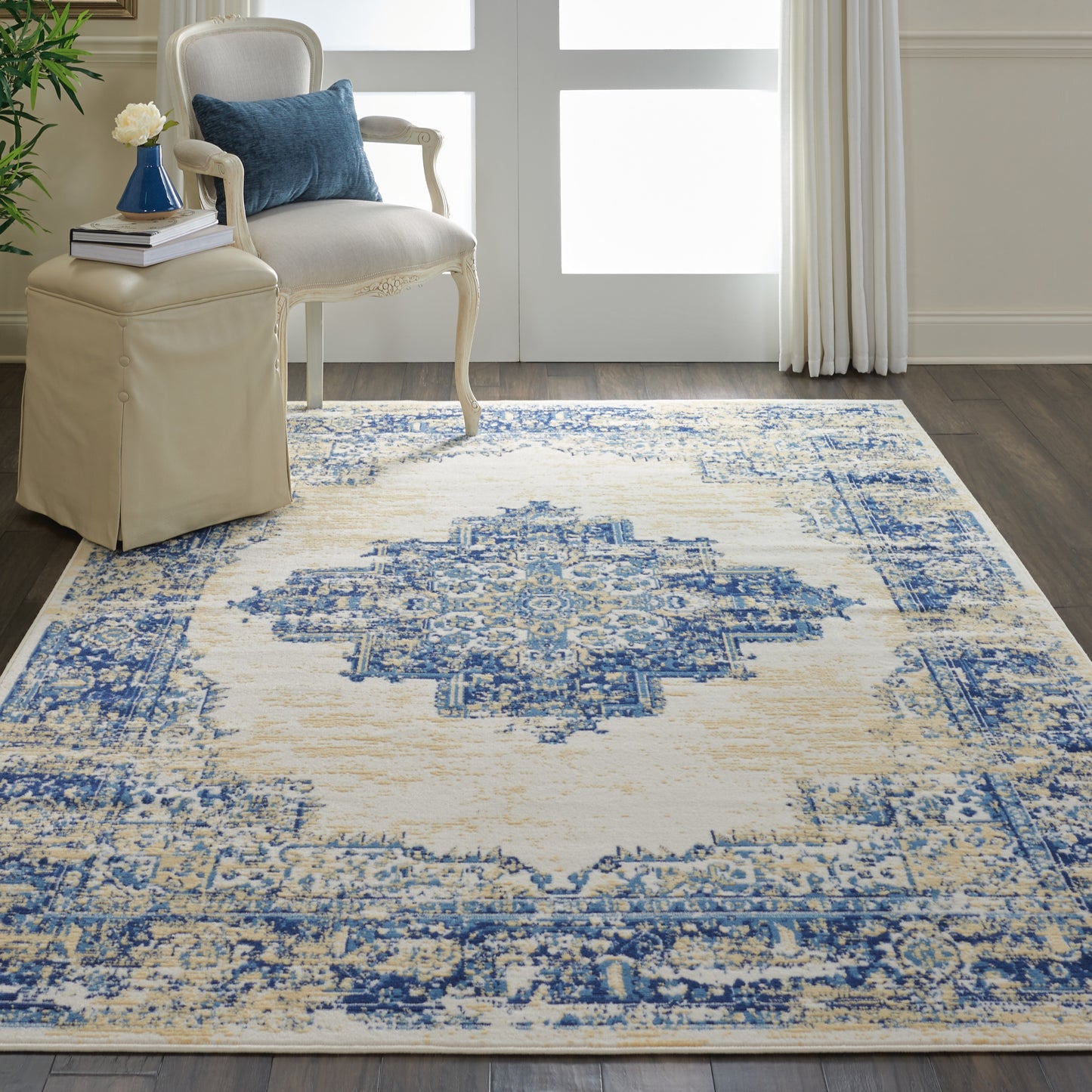 Blue and ivory distressed oriental rug with central medallion on dark wood floor; cream carved-armchair, blue cushion, beige ottoman.