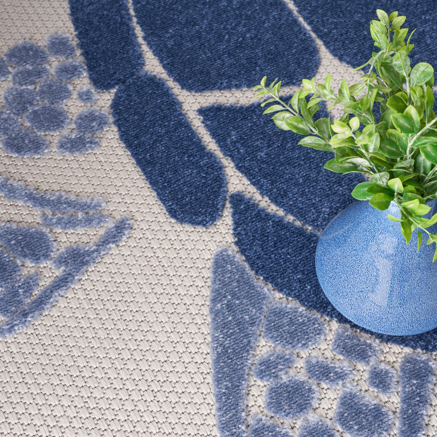 Beige & blue patterned woven rug with a blue ceramic vase & green leafy plant.