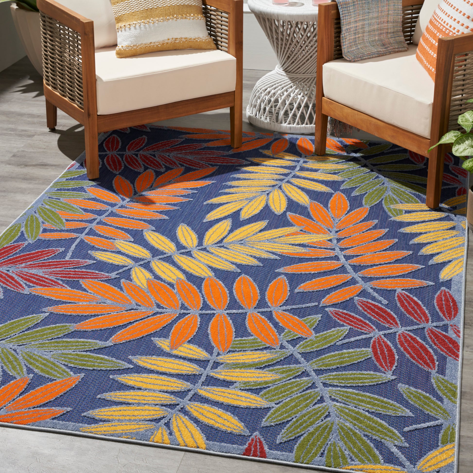 Blue outdoor rug with vivid multicolor leaf pattern (orange, yellow, green, red) under wooden-framed chairs.