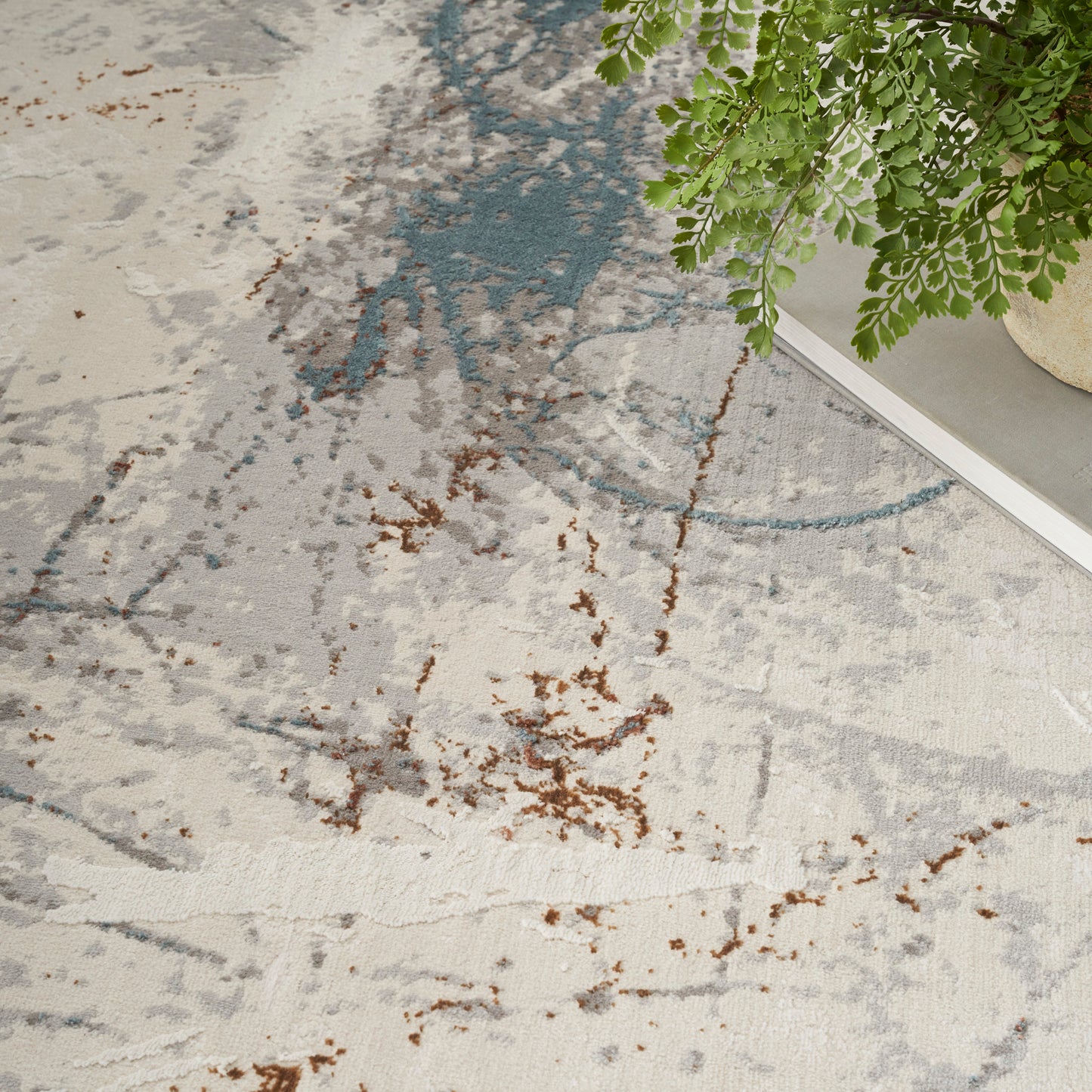 Modern abstract area rug in beige & gray with blue & brown splatters, paired with a green leafy plant.